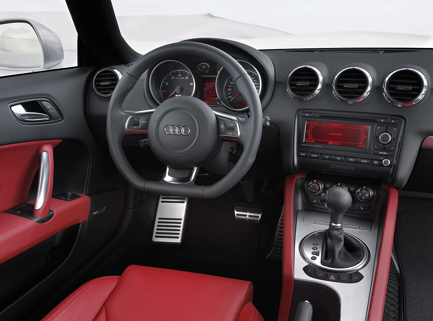 Innenraum eines Audi mit schwarzem Lenkrad, roten Ledersitzen und modernen Armaturen. Manuelle Gangschaltung und silberne Pedale.
