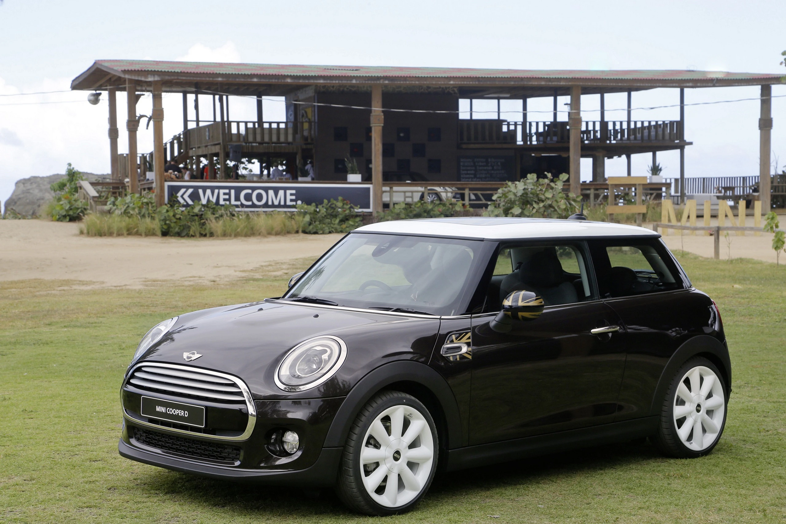 Schwarzer Mini Cooper mit weißen Felgen steht auf einer Wiese. Im Hintergrund befindet sich ein großes Gebäude mit einem "Welcome"-Schild.