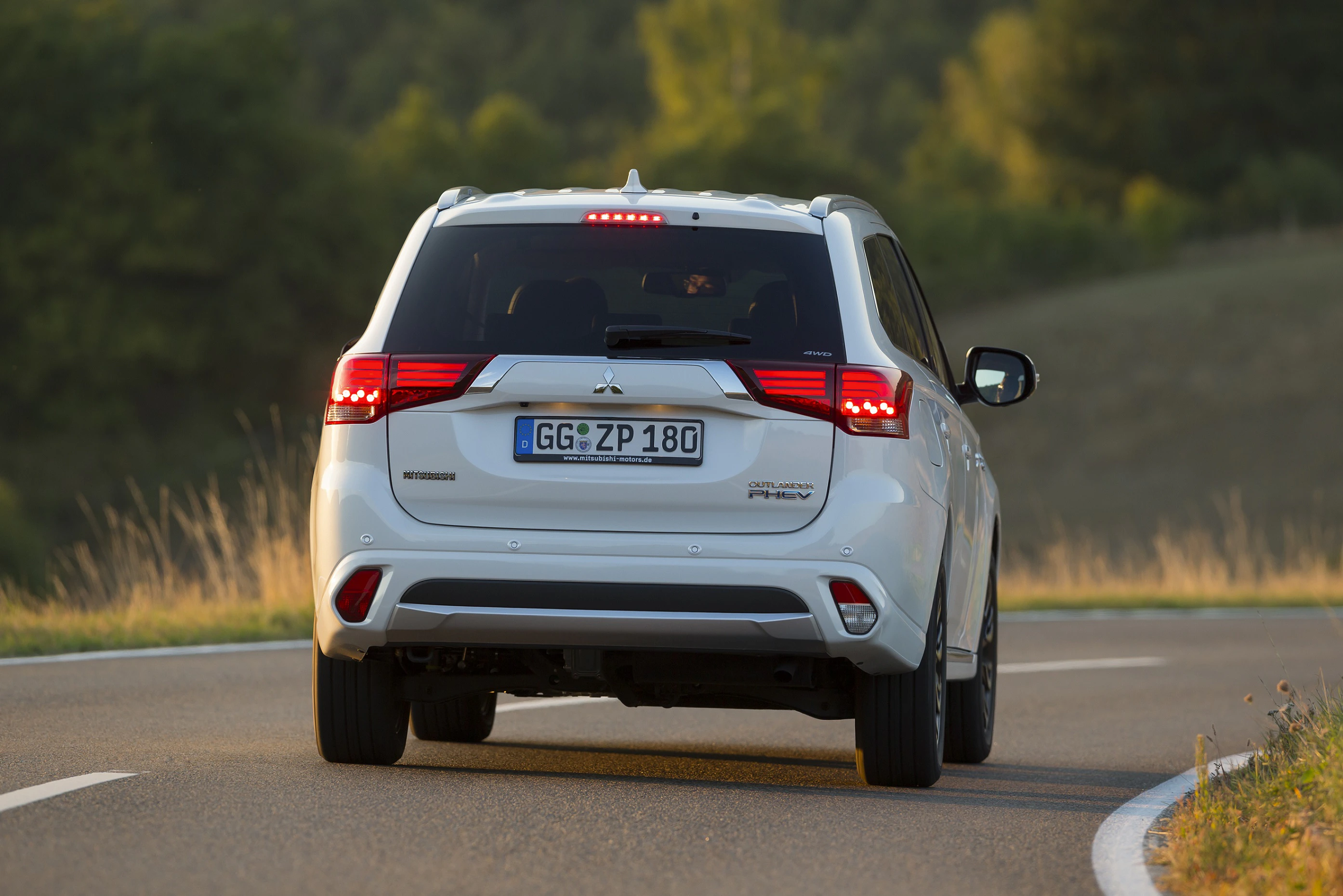 Weißer SUV fährt auf einer kurvigen Landstraße, umgeben von grüner Landschaft und Wald im Hintergrund.