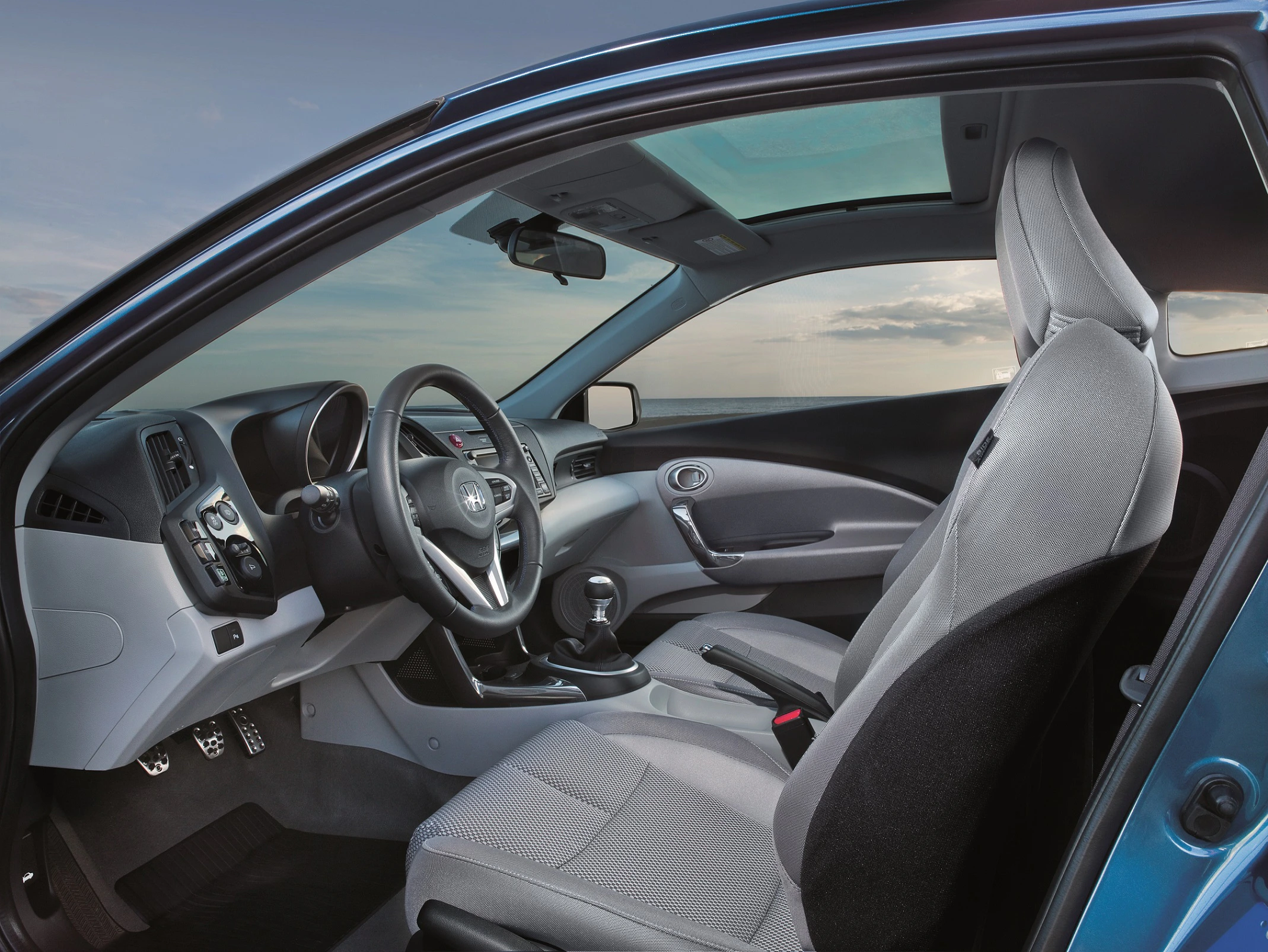 Innenraum eines modernen Autos mit hellgrauen Sitzen, großzügigem Glasdach und Armaturenbrett in dunklen Tönen. Fahrzeug steht vor einem blauen Himmel.