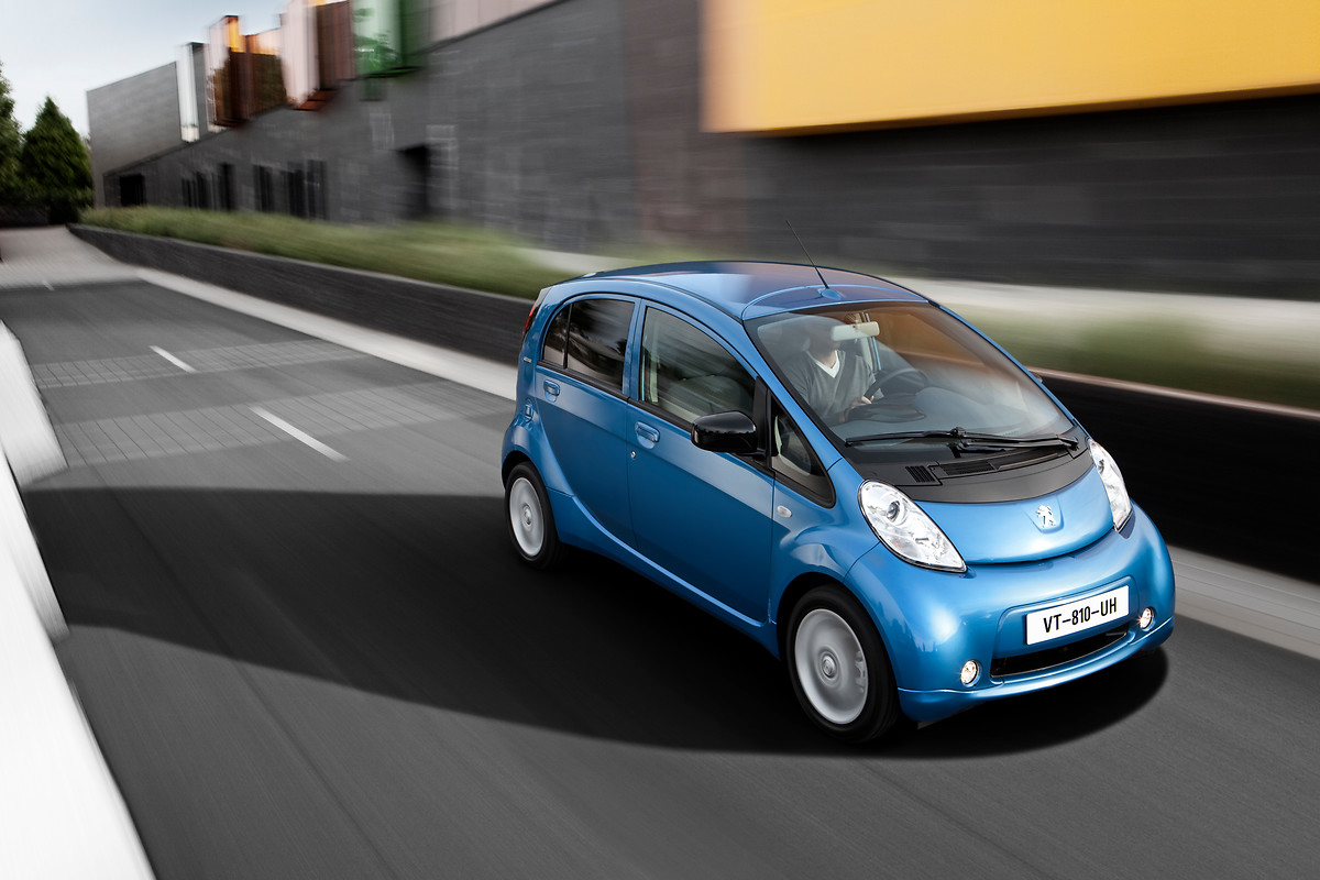 Blaues Elektroauto fährt auf städtischer Straße an modernen Gebäuden vorbei. Fahrer im Auto, klare Himmel und stilvolle Architektur.