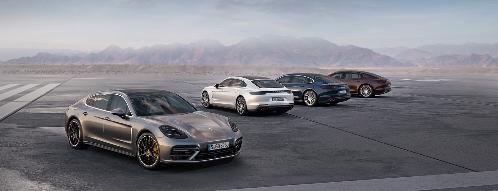 Vier sportliche Autos stehen auf einer großen Freifläche vor einer Bergkulisse unter klarem Himmel.