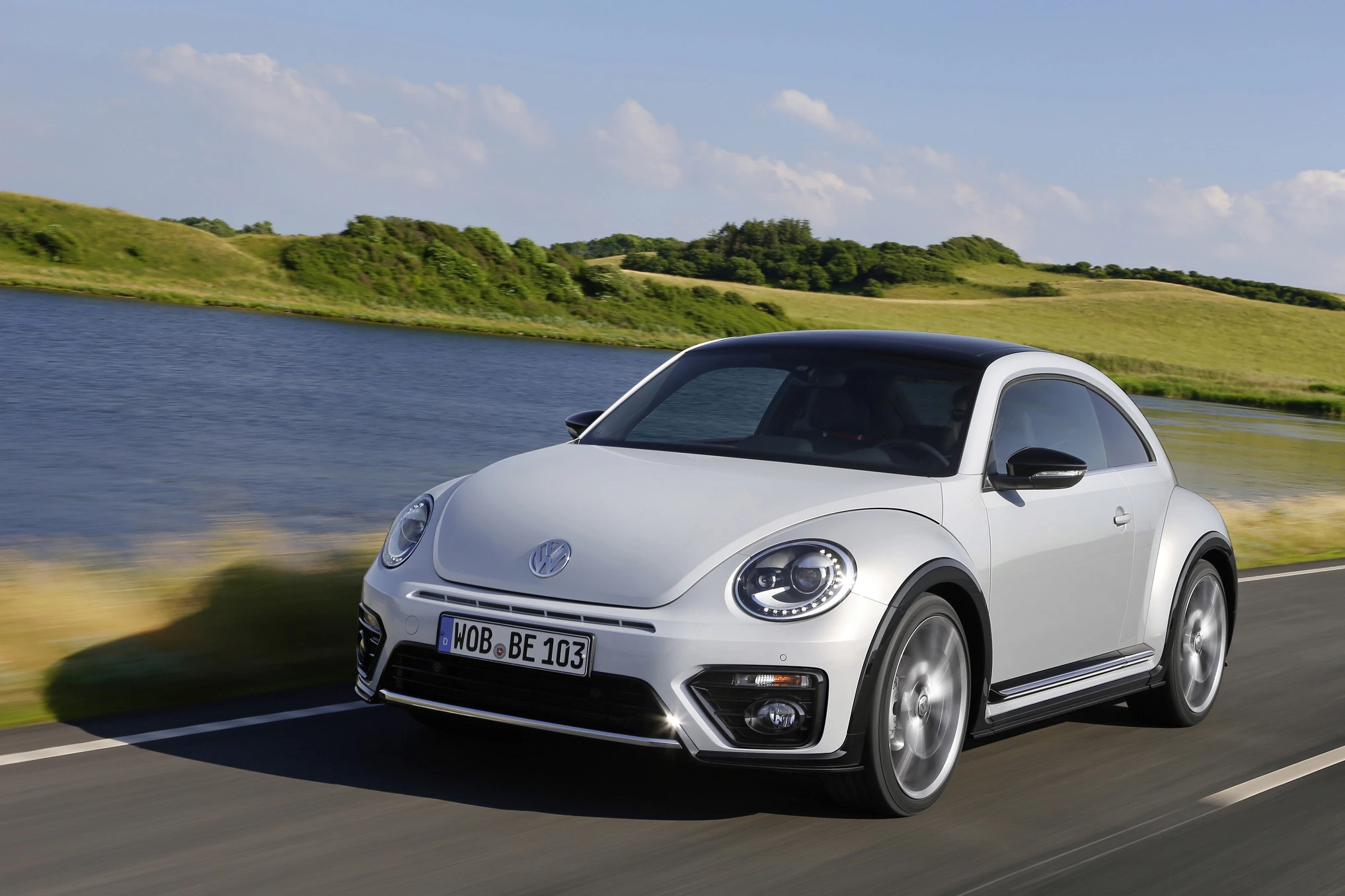 Ein weißer VW Beetle fährt auf einer Landstraße entlang eines Sees, umgeben von grünen Hügeln unter blauem Himmel.