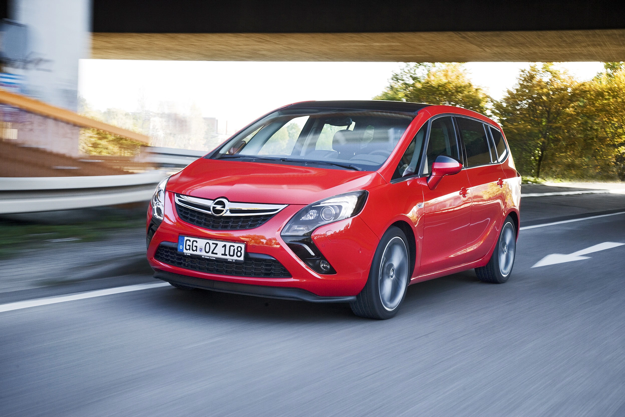 Roter Opel Zafira Tourer fährt auf einer Straße unter einer Brücke entlang, Hintergrund verschwommen.