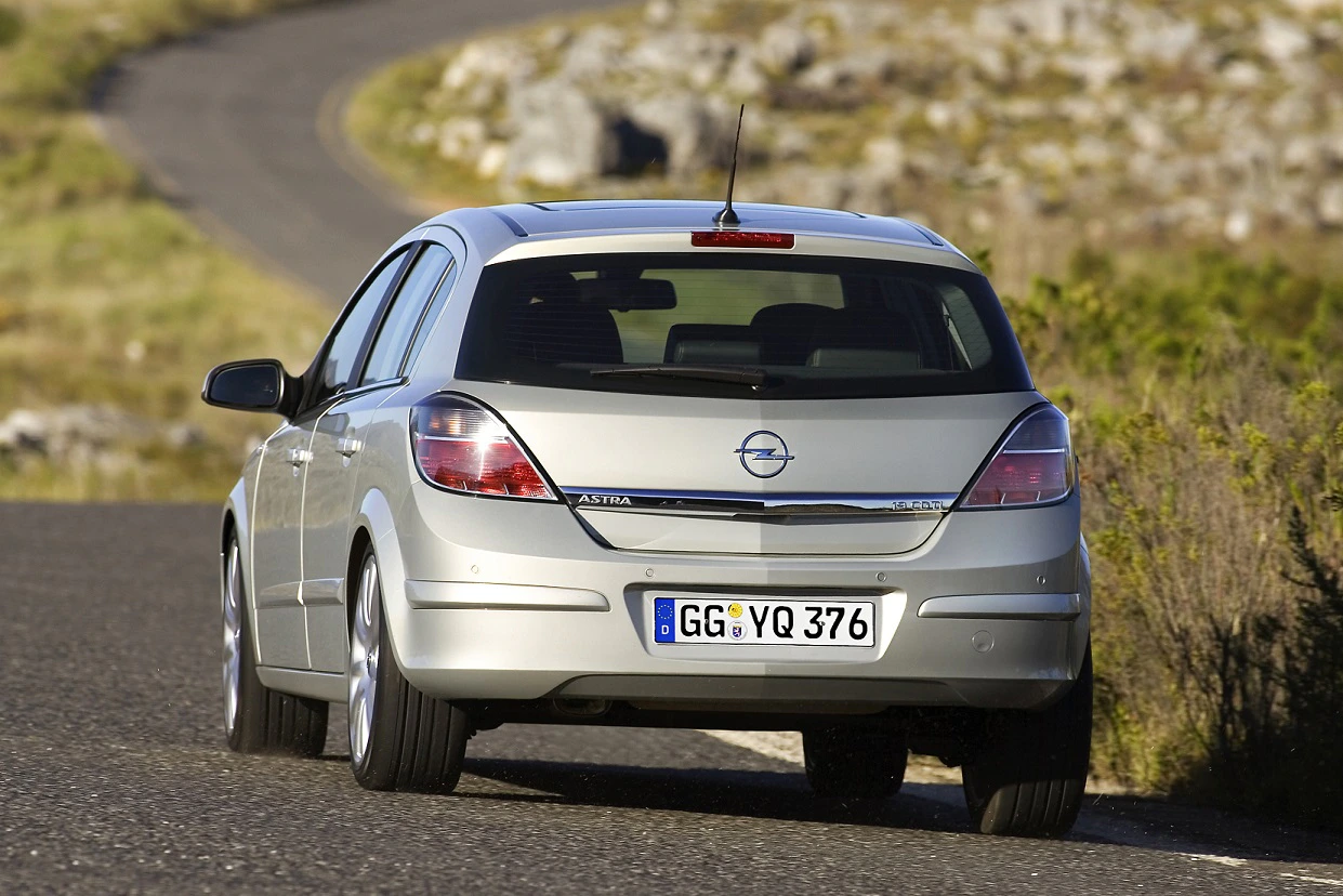 Silberner Opel Astra fährt auf einer kurvigen Landstraße. Im Hintergrund sind Pflanzen und ein felsiger Hügel zu sehen.