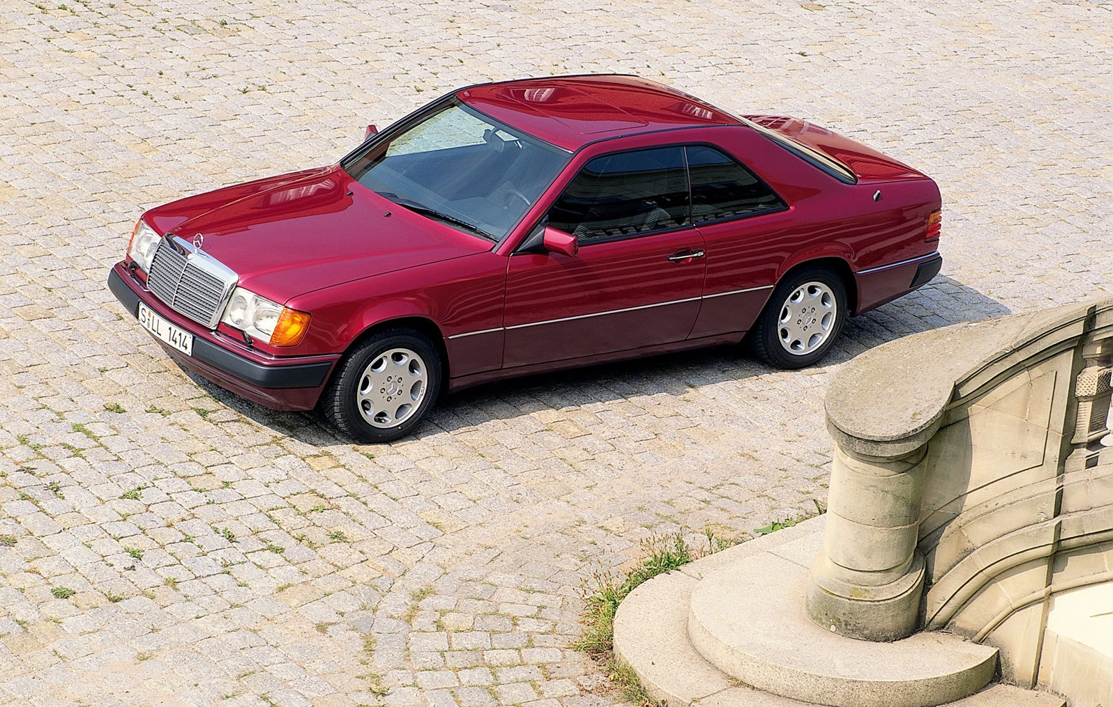 Rotes Coupé auf Kopfsteinpflaster, seitlich von oben fotografiert. Elegantes Design, klare Linien, parkend neben steinernen Stufen.