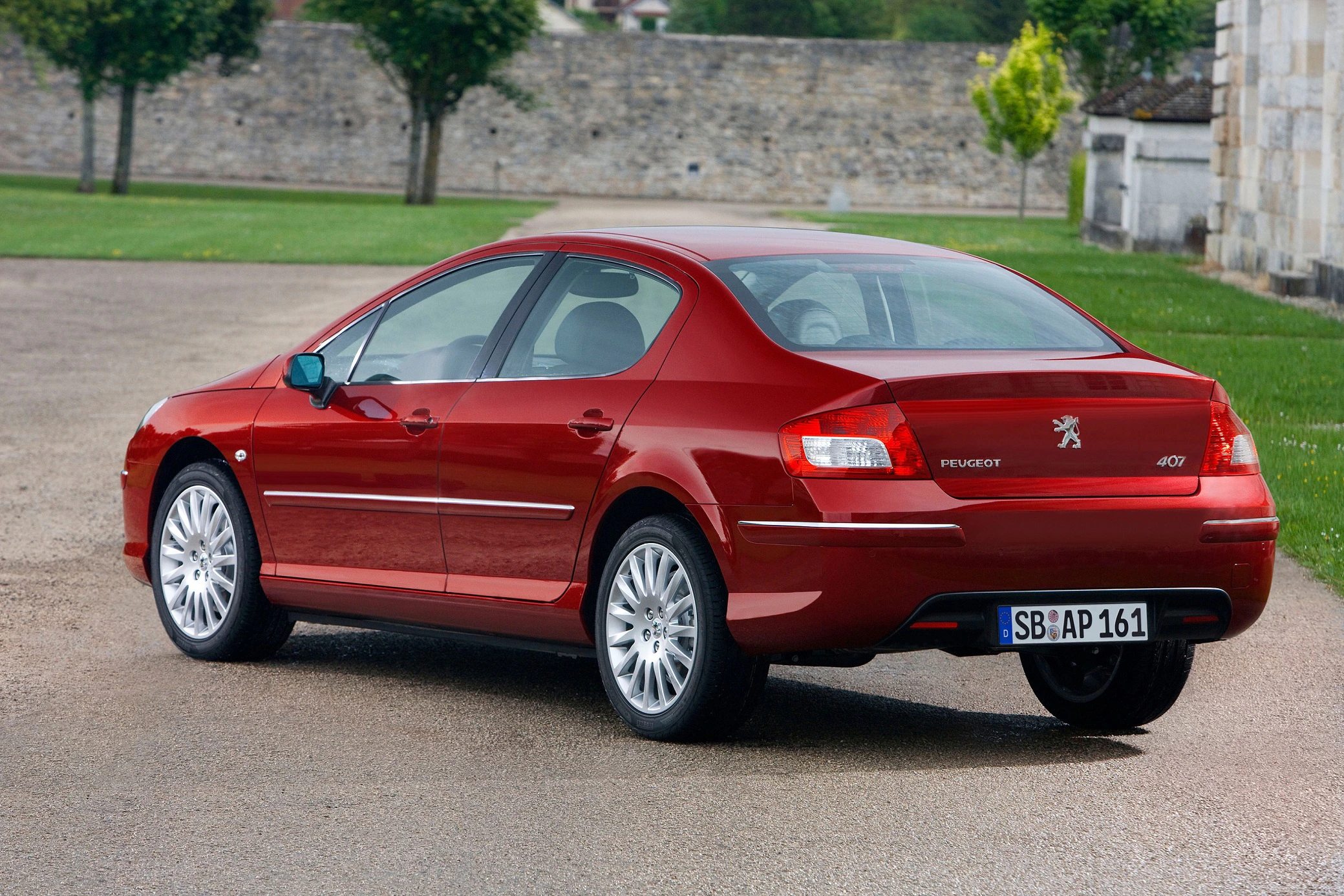 Ein roter Peugeot 407 steht auf einer Straße im Freien, umgeben von Grünflächen und einer Mauer im Hintergrund.