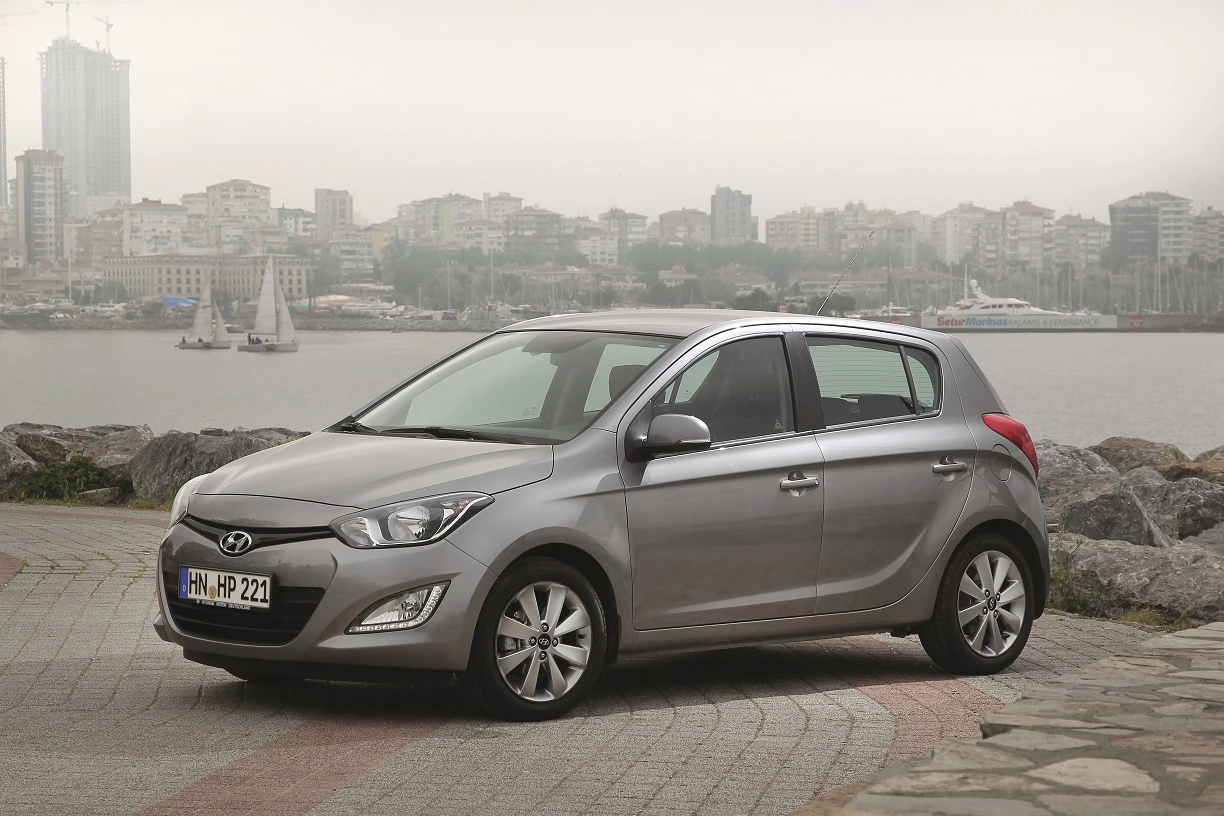 Silberner Hyundai i20 steht auf einer gepflasterten Fläche vor einer nebligen Stadtlandschaft mit Segelbooten und modernen Gebäuden im Hintergrund.