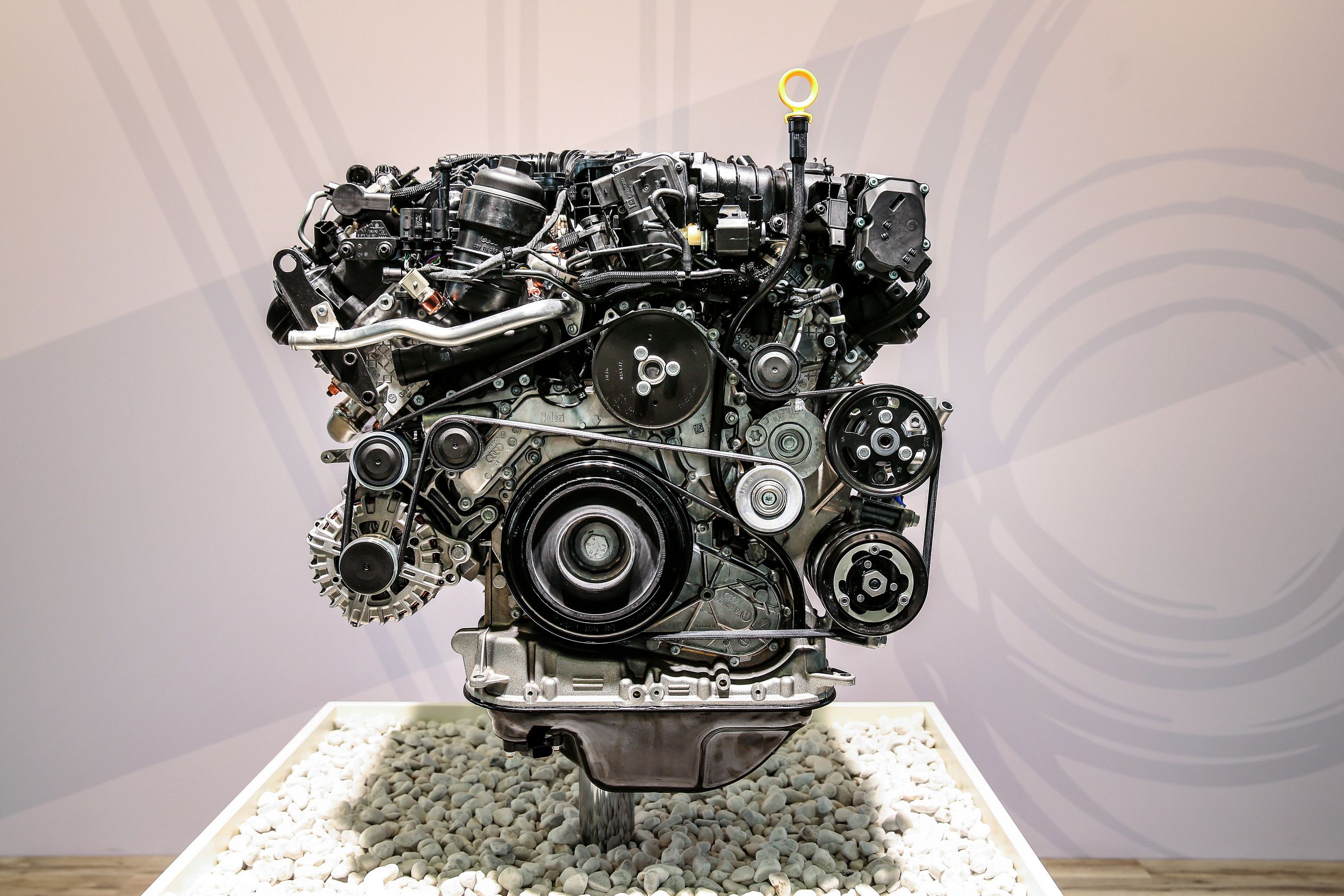 Nahaufnahme eines modernen Automotors auf einem Podest, umgeben von hellen Steinen, zeigt verschiedene Riemen und mechanische Teile.