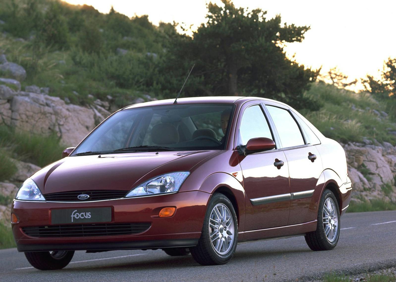 Roter Ford Focus Limousine parkt auf einer Landstraße, umgeben von grünen Bäumen und Felsen im Hintergrund.