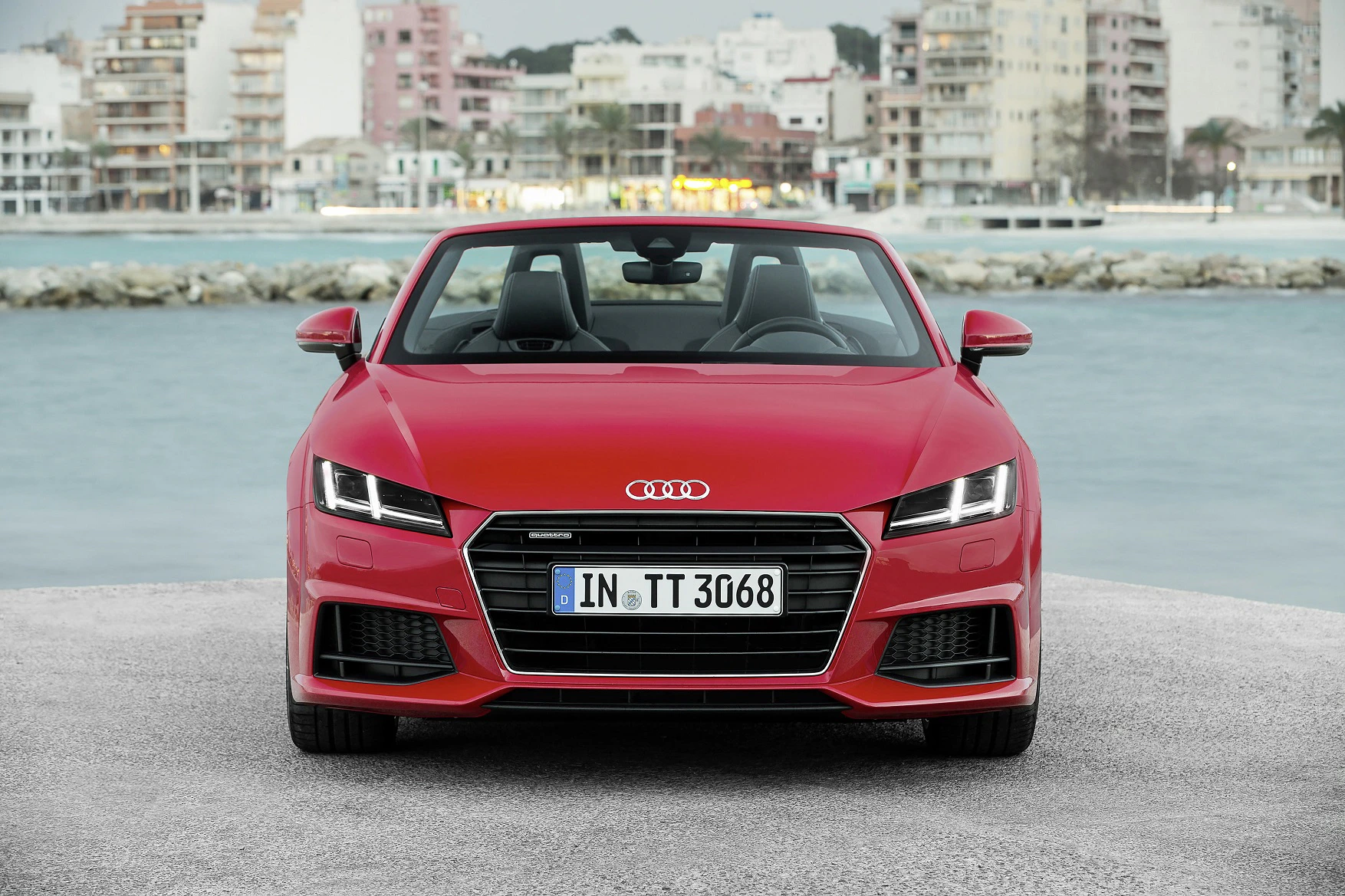Roter Audi TT Cabrio mit geöffnetem Verdeck steht vor einer Küstenkulisse, moderne Gebäude im Hintergrund.