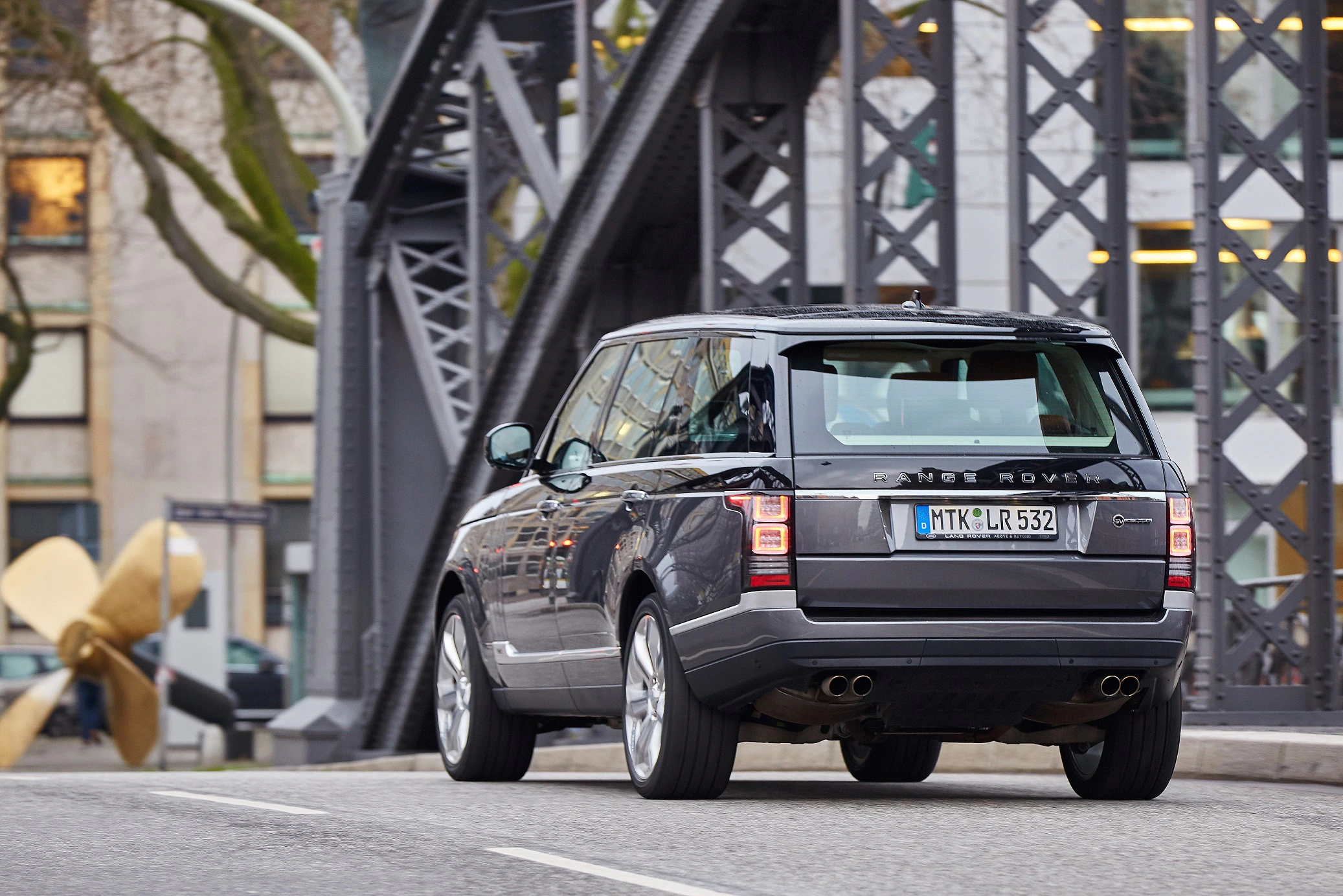 Ein schwarzer Range Rover fährt über eine Brücke in einer Stadtumgebung. Im Hintergrund sind Gebäudefassaden und ein großes Schraubenmodell zu sehen.
