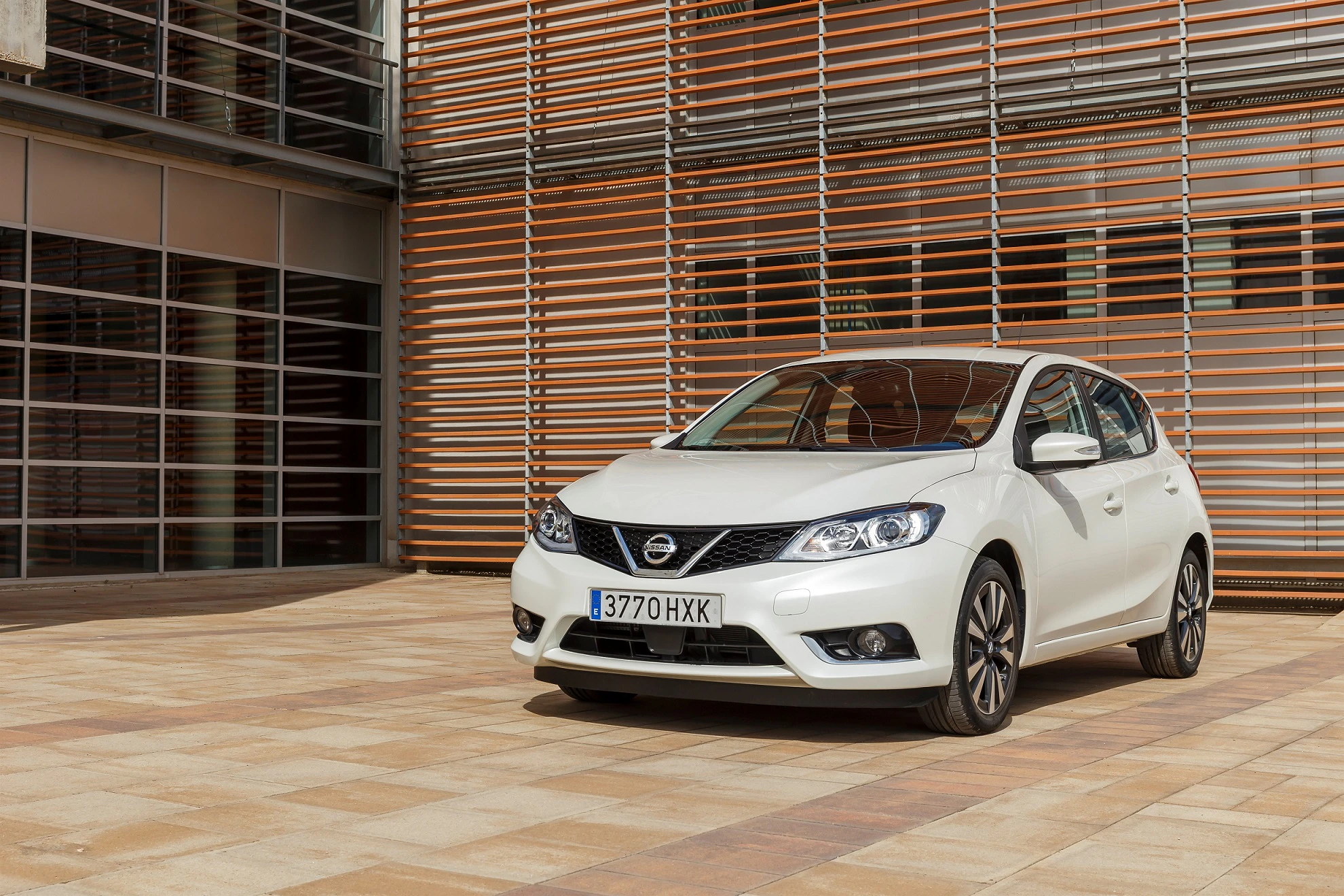 Weißer Nissan Pulsar steht auf einem gepflasterten Platz vor einem modernen Gebäude mit Holzlamellen-Fassade.