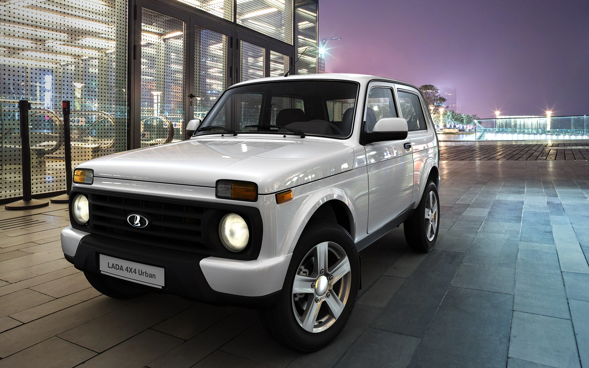Weißer Lada 4x4 Urban steht nachts beleuchtet vor einem modernen Gebäude mit Glasfassade, Scheinwerfer eingeschaltet.
