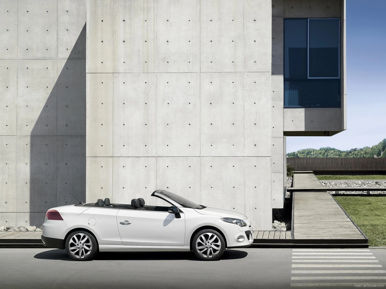 Weißes Cabriolet parkt vor einer modernen Betonwand. Rechts ist ein Gebäude mit großen Glasfenstern und eine Terrasse mit Steinen zu sehen.