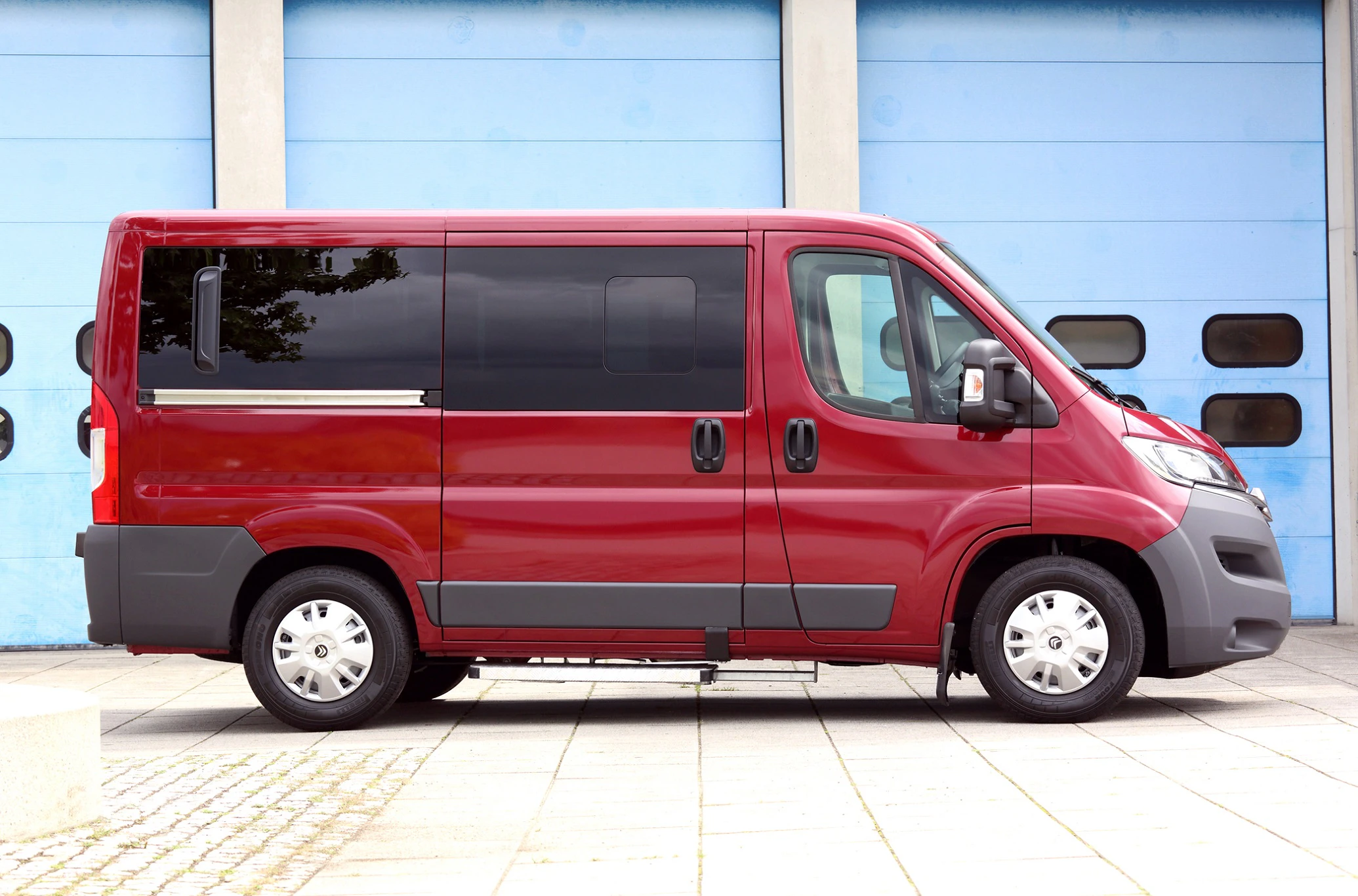 Roter Kleinbus vor blauer Garage, seitlich aufgenommen. Fahrzeug mit dunklen Fenstern und drei Türen sichtbar.