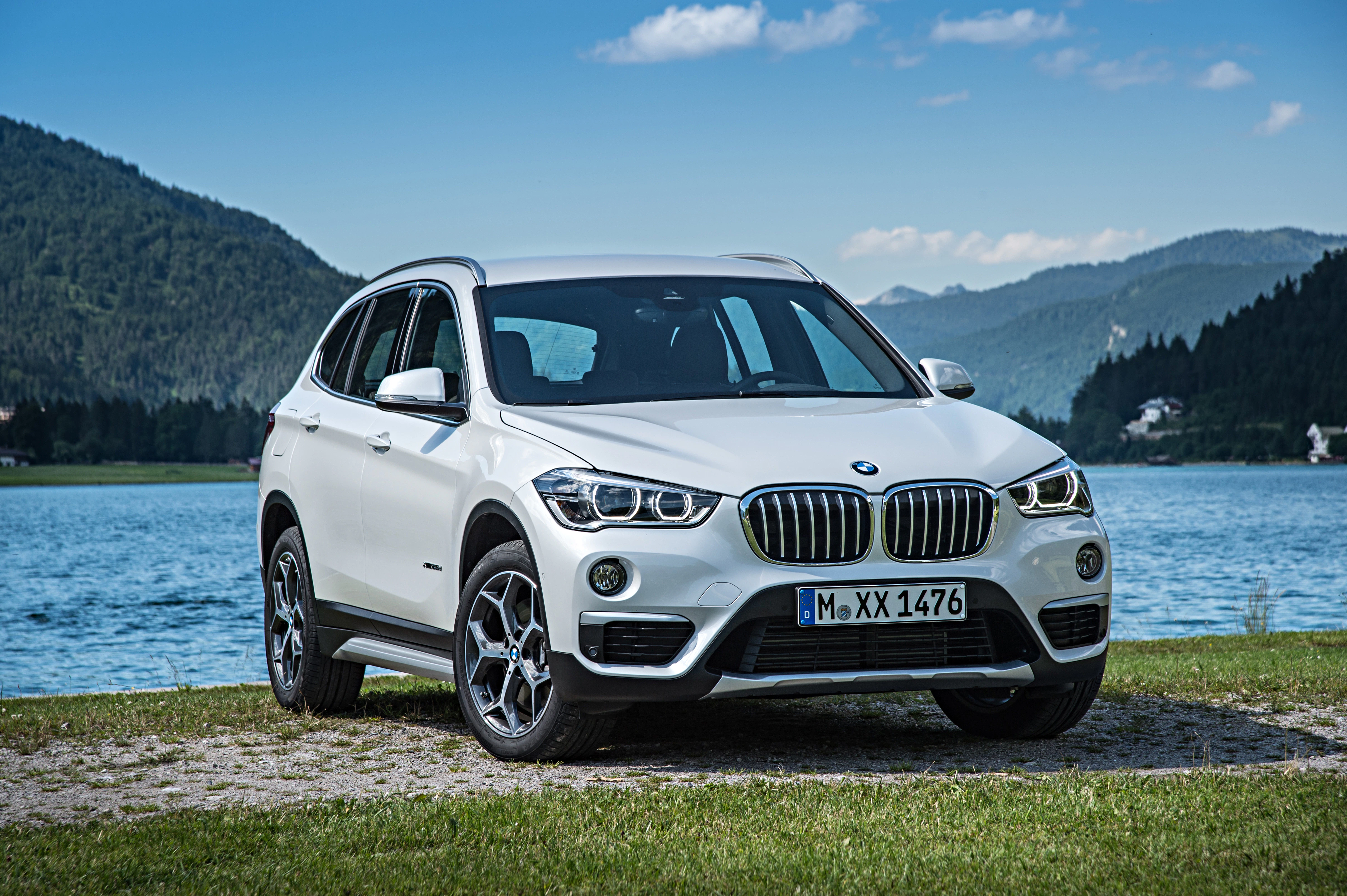 Weißer SUV parkt vor einem malerischen See und bewaldeten Bergen bei blauem Himmel.
