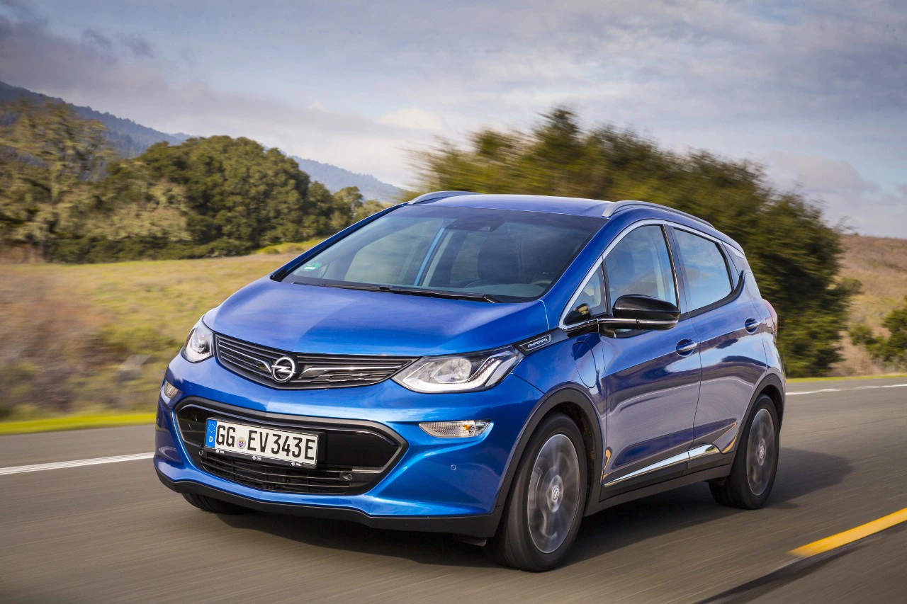 Blauer Opel Ampera-e fährt auf einer Landstraße, umgeben von grüner Landschaft und bewölktem Himmel.