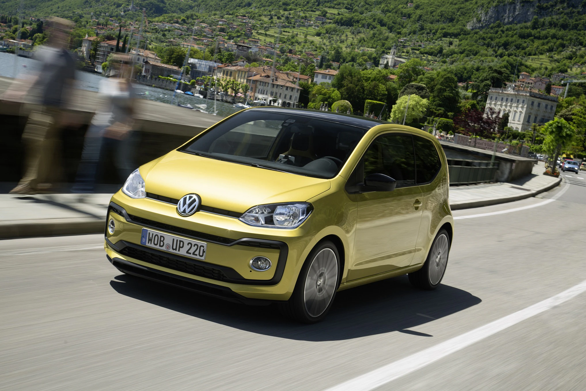 Gelber VW up! fährt auf einer malerischen Straße durch eine grüne, hügelige Landschaft mit verschwommenen Fußgängern im Hintergrund.