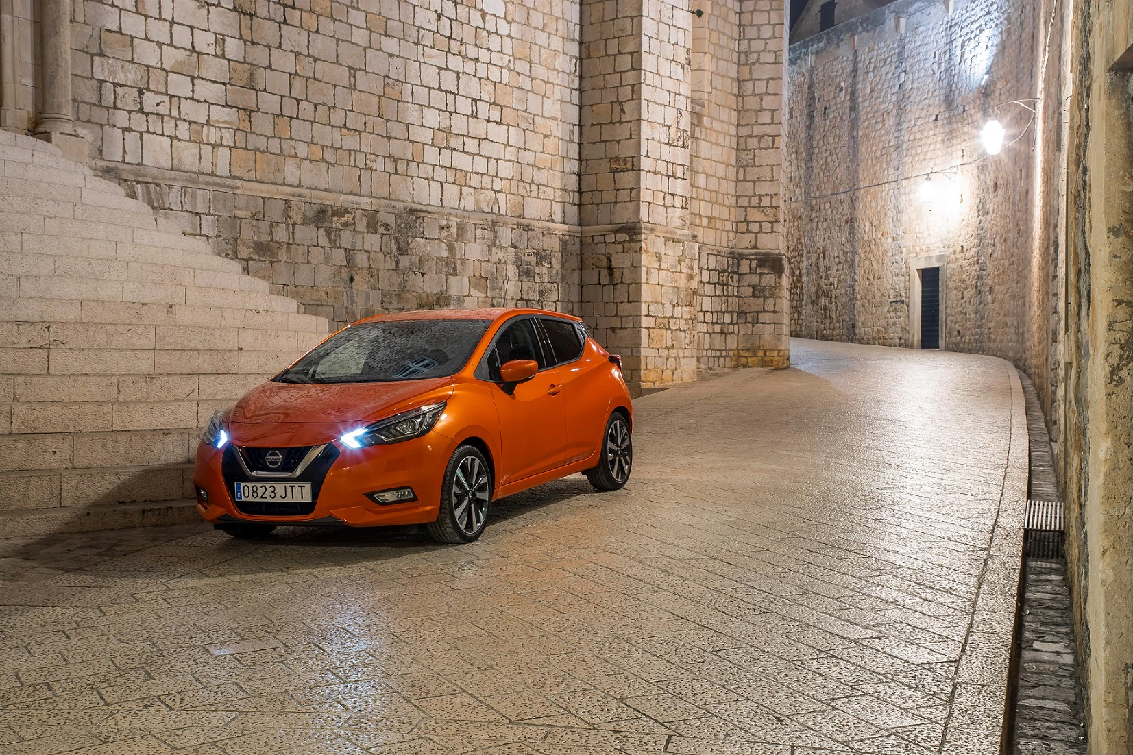 Ein orangefarbener Nissan-Fahrzeug steht auf einer gepflasterten Straße, umgeben von hohen, steinernen Mauern einer Altstadt bei Nacht.