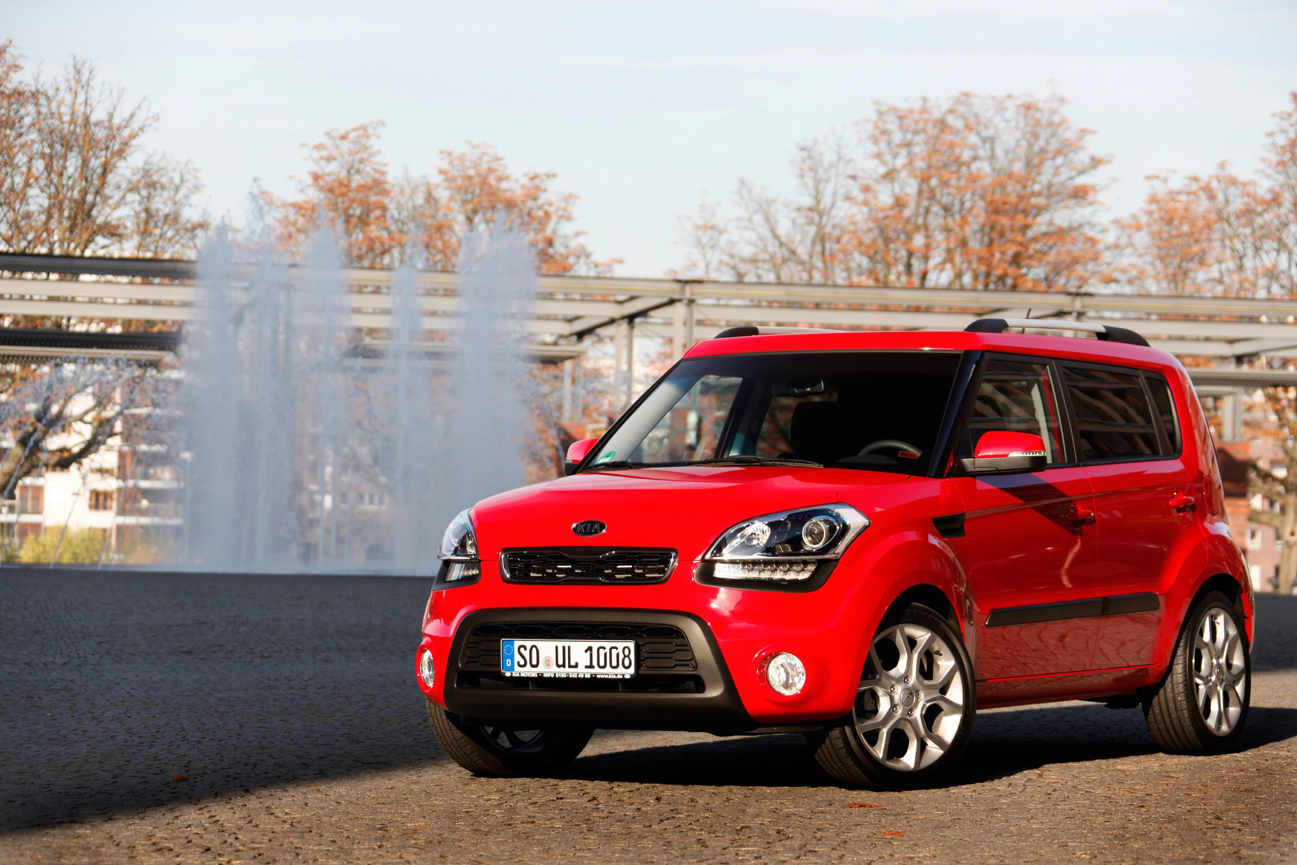 Roter SUV parkt vor einem sprühenden Brunnen, umgeben von kahlen Bäumen unter klarem Himmel.