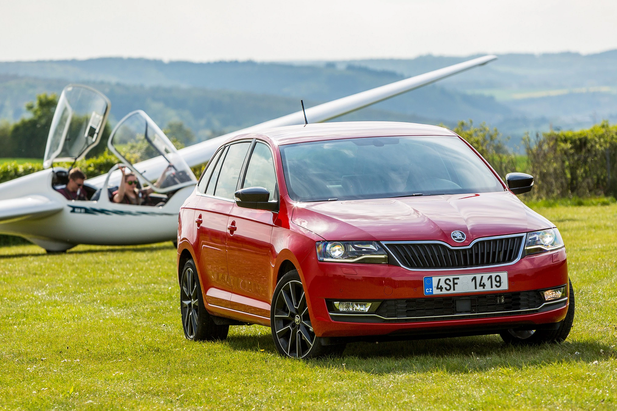 Roter Skoda auf einer Wiese, im Hintergrund ein Segelflugzeug und zwei Personen, die sich zum Start vorbereiten. Hügelige Landschaft im Hintergrund.