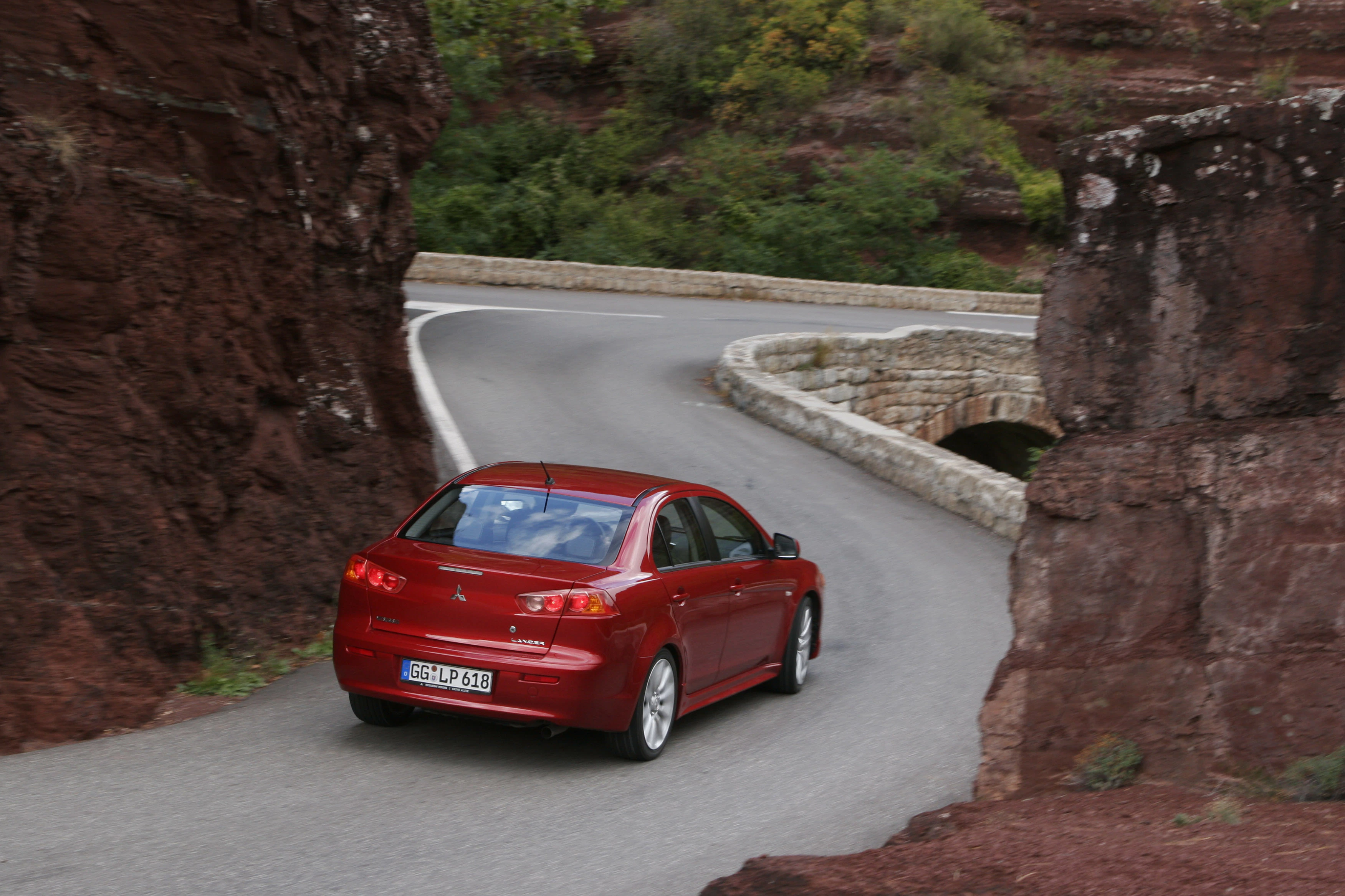 Rotes Auto fährt auf schmaler, kurviger Bergstraße zwischen felsigen Klippen und grünem Bewuchs.