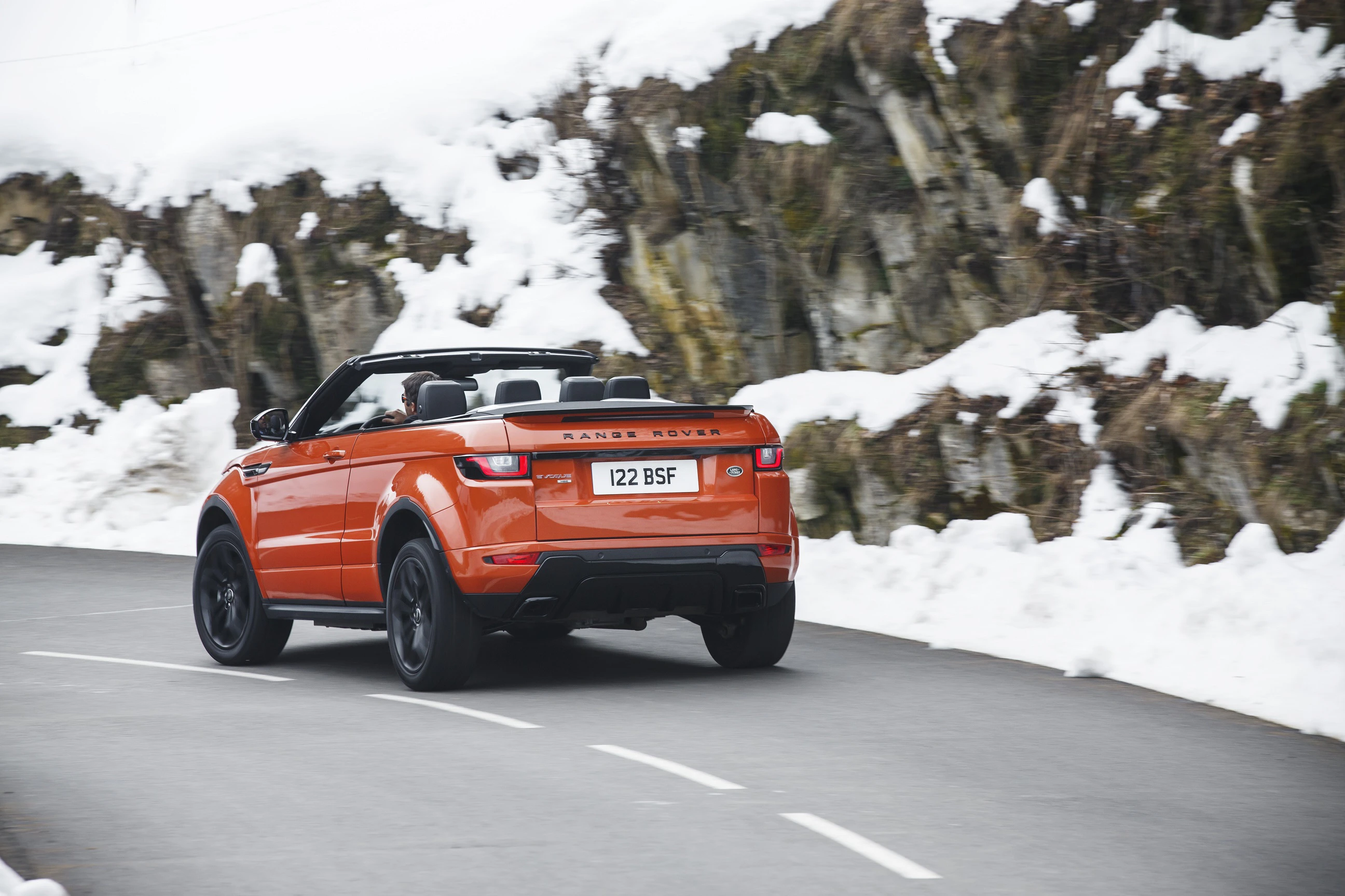 Ein orangefarbener Cabrio-Geländewagen fährt auf einer verschneiten Bergstraße entlang einer Felswand.