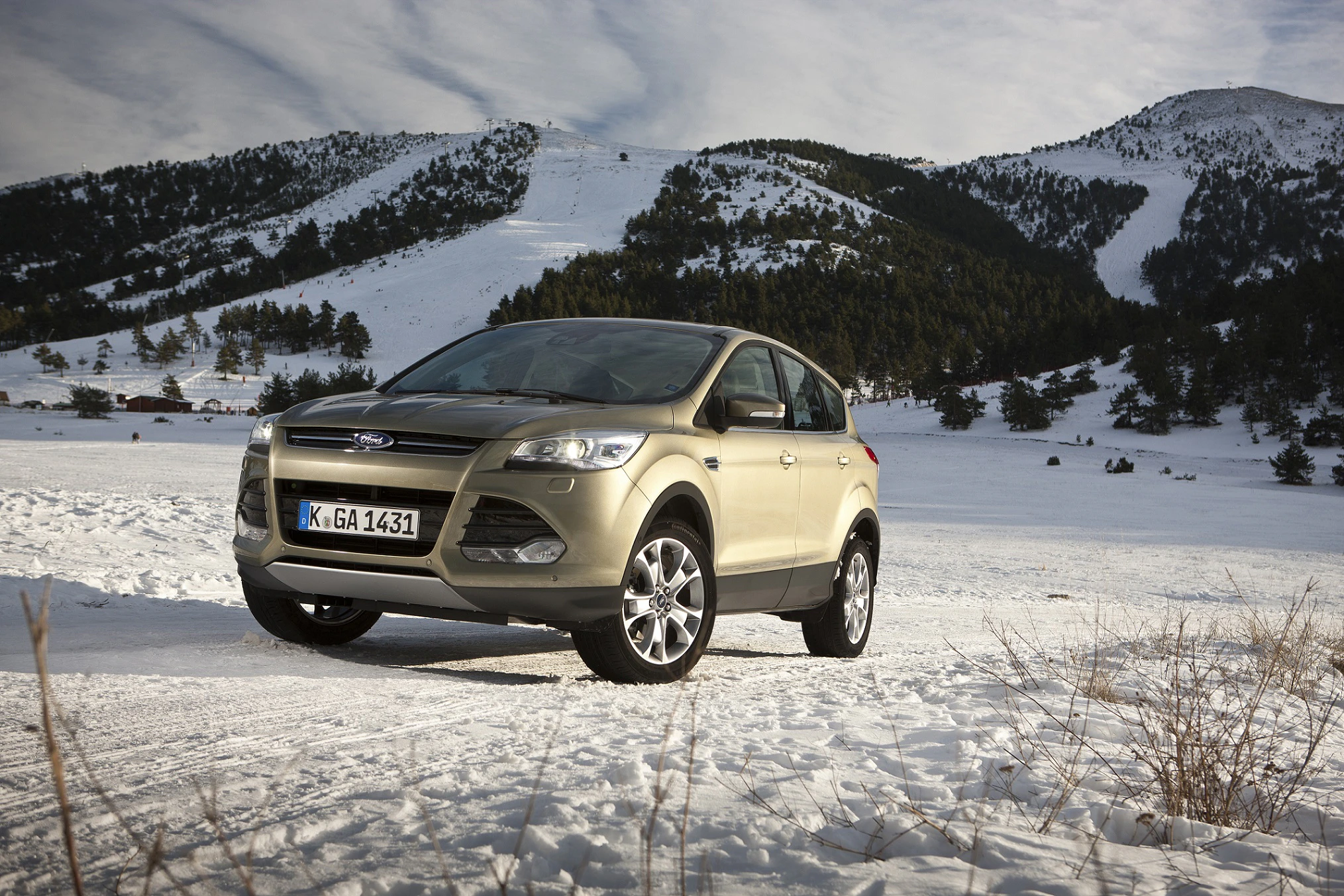 Ein beigefarbener SUV fährt auf einem verschneiten Weg vor einer Berglandschaft mit bewaldeten Hängen und blauem Himmel.