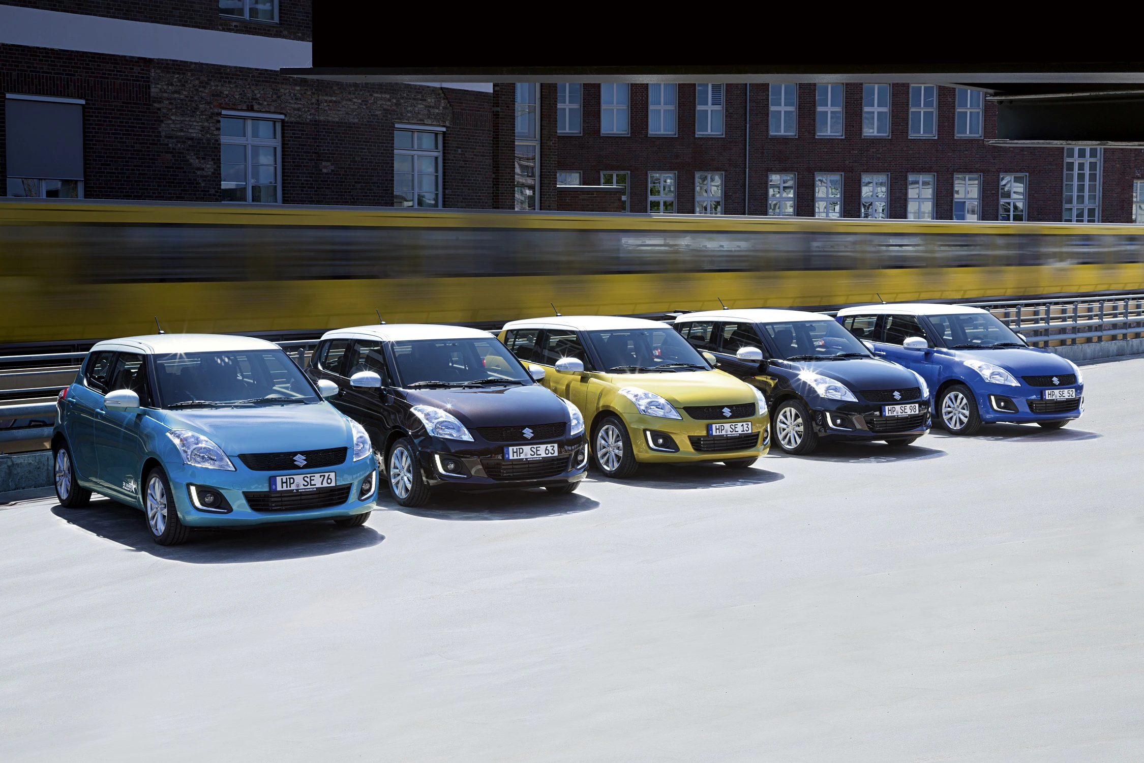 Sechs bunte Autos, darunter blau, schwarz und gelb, parken nebeneinander vor einem verschwommenen vorbeifahrenden Zug im Hintergrund.