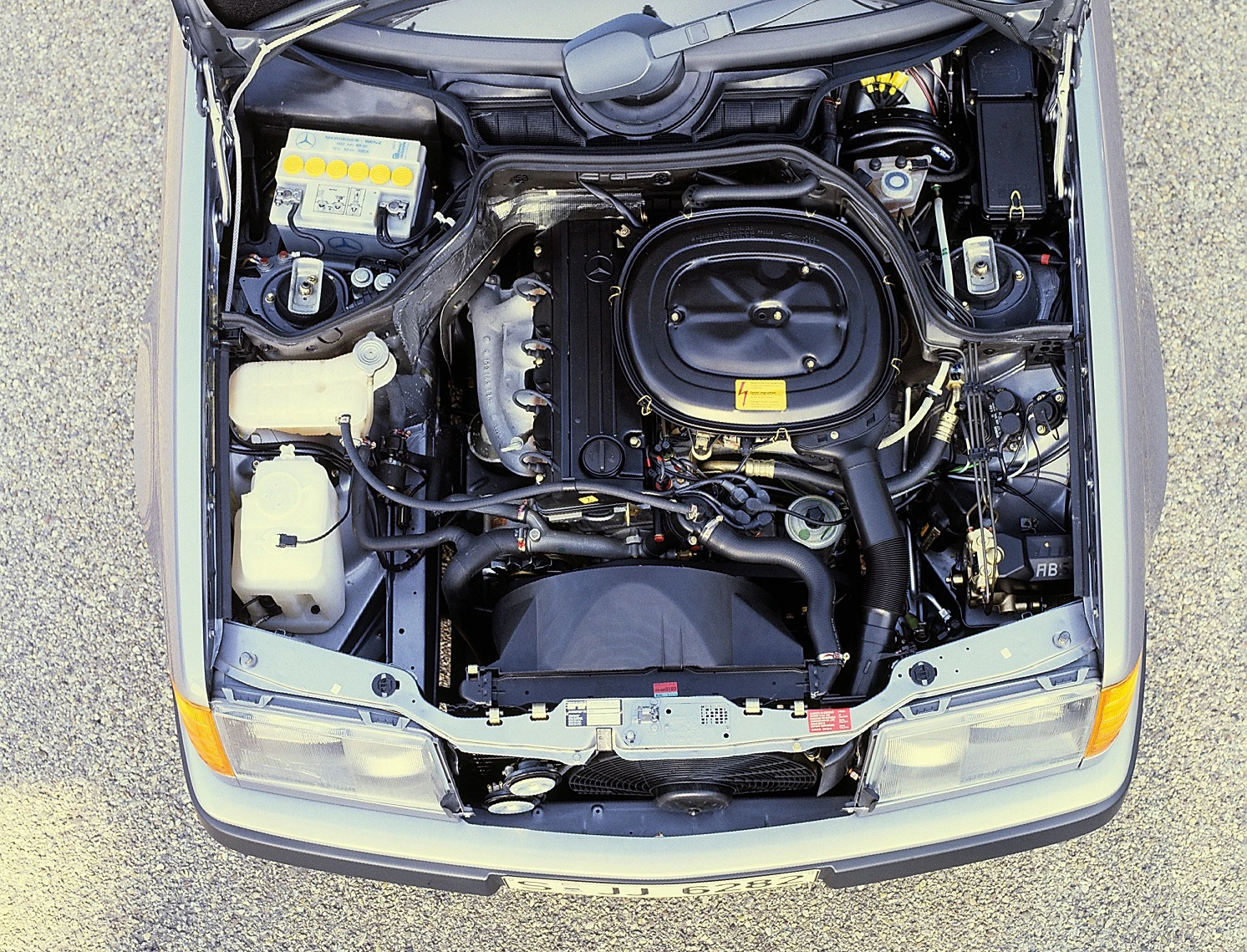Motorraum eines Autos von oben, mit sichtbarem Motorblock, Batterie, Kühlmittelbehälter und anderen mechanischen Komponenten.