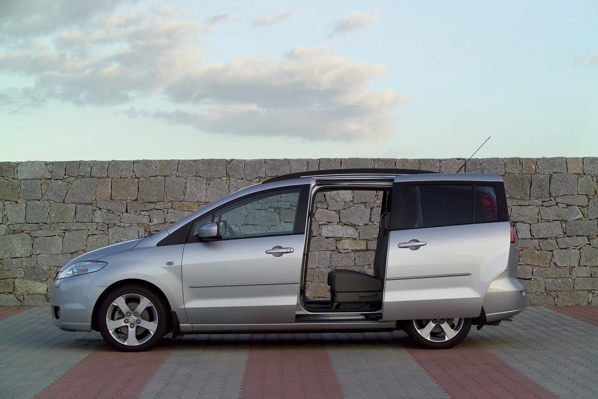 Silberner Minivan mit geöffneten Seitentüren, geparkt vor einer Steinmauer unter einem Himmel mit vereinzelten Wolken.
