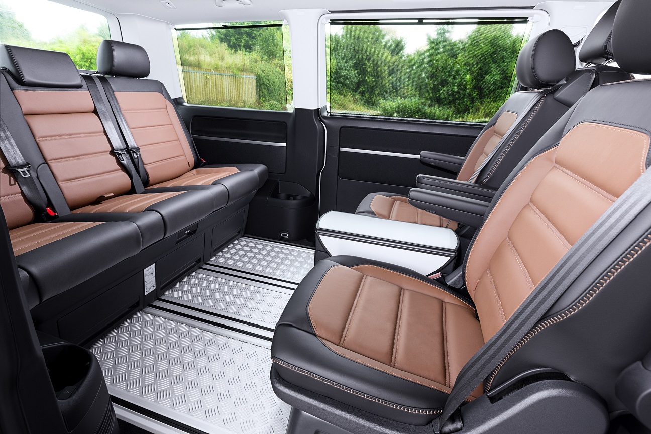 Innenraum eines Transporters mit vier braunen Ledersitzen, zwei vordere um 180° gedreht, und einem Rücksitz, sichtbar sind grüne Bäume durch die Fenster.