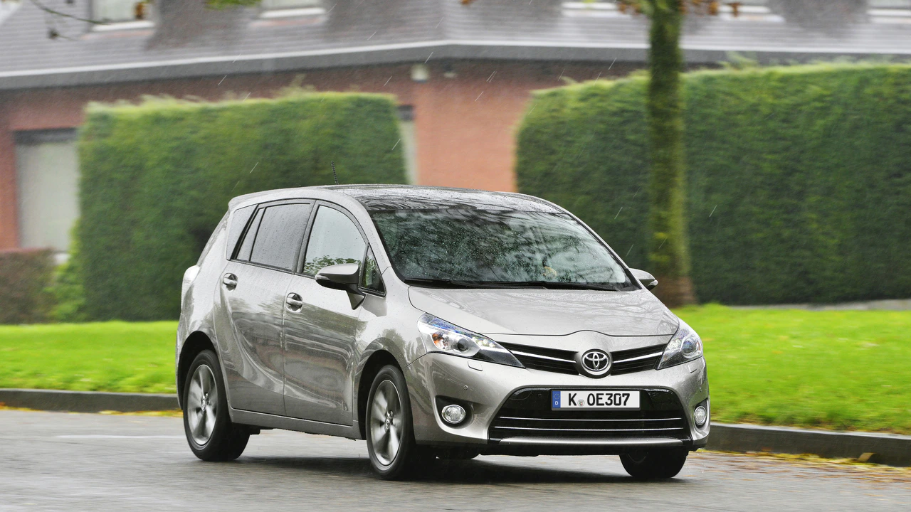 Silberner Toyota Kleinwagen fährt bei Regen auf einer Straße entlang grüner Hecken und Ziegelgebäude im Hintergrund.