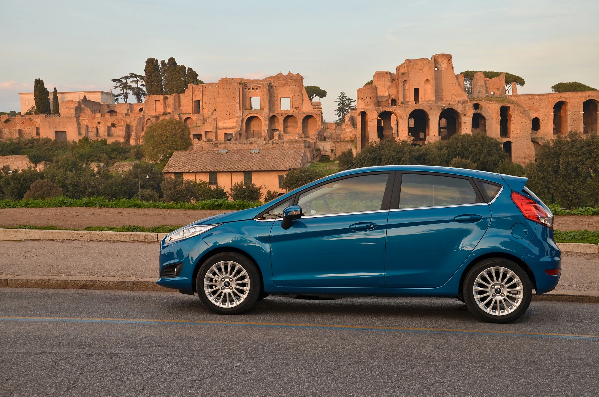 Blaues Auto, geparkt vor antiken römischen Ruinen bei Sonnenuntergang, mit Bäumen und einem klaren Himmel im Hintergrund.
