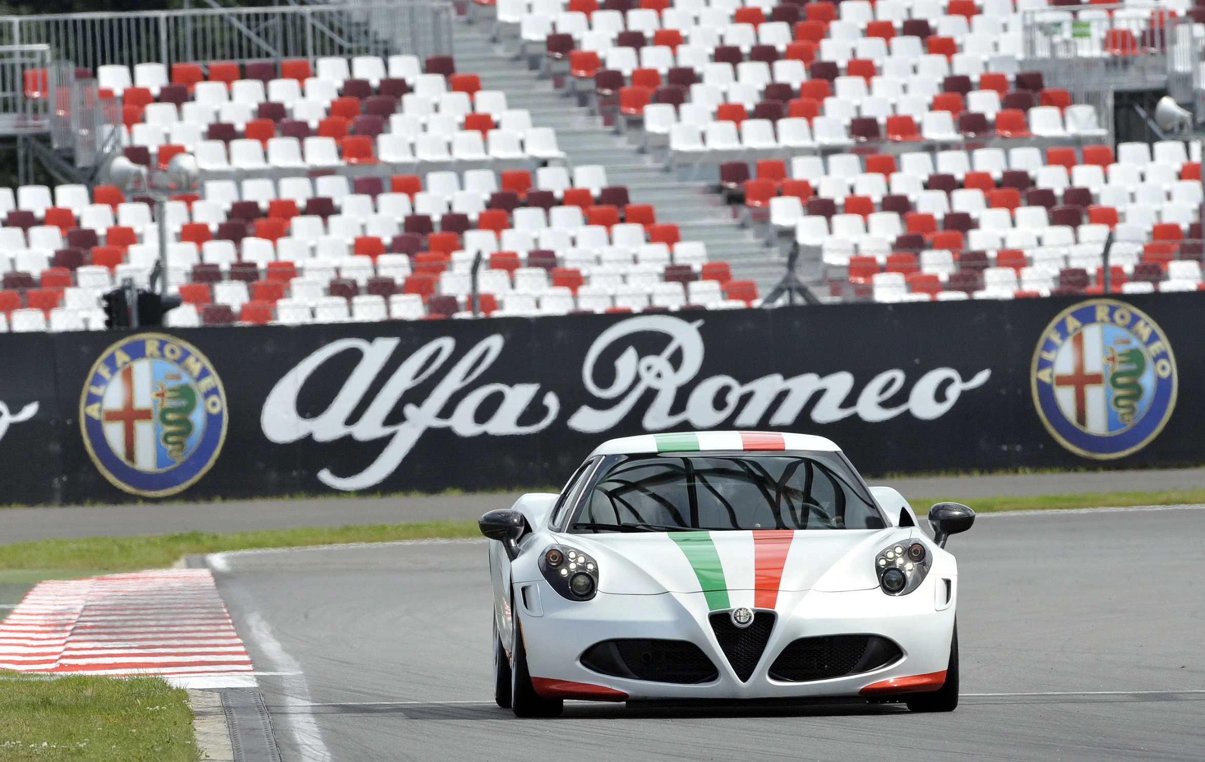 Ein weißer Alfa Romeo Sportwagen mit italienischen Streifen fährt auf einer Rennstrecke, im Hintergrund leere Tribünen.