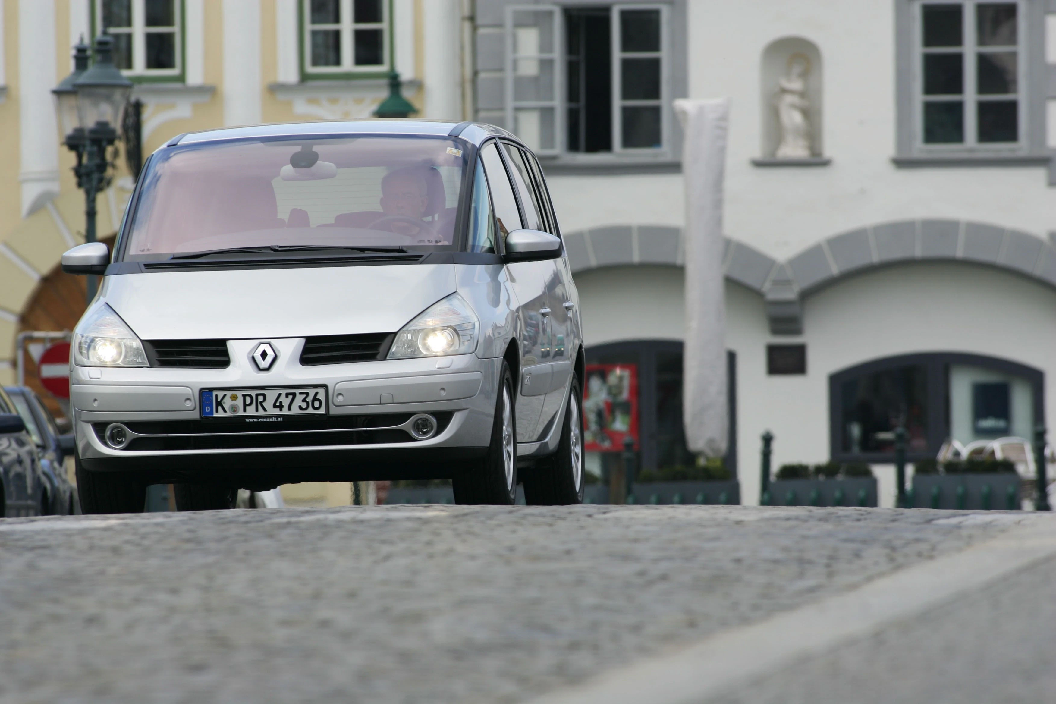 Silberner Renault Scenic fährt auf Kopfsteinpflasterstraße in einer Altstadt mit historischen Gebäuden im Hintergrund.