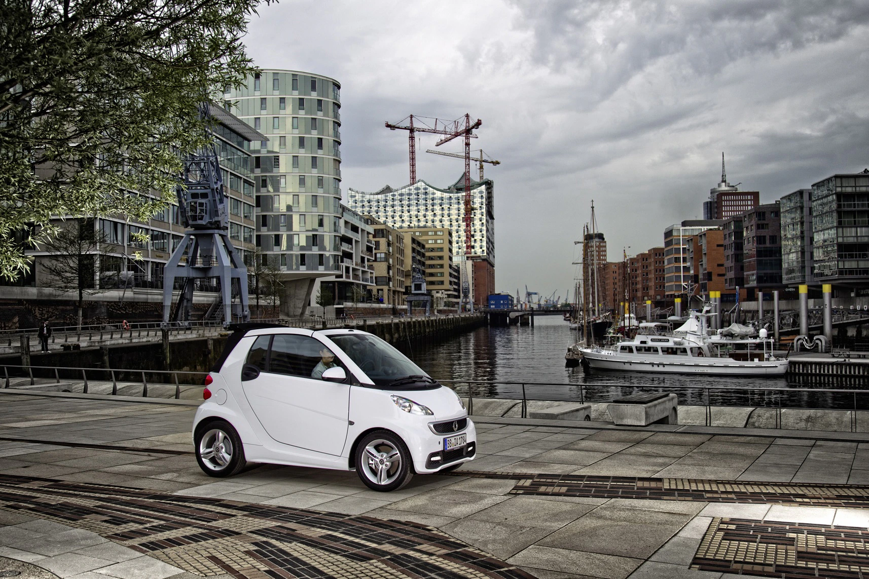 Ein weißer Smart steht in einer modernen Stadtlandschaft am Wasser, mit Booten und im Hintergrund die Elbphilharmonie und Baukräne.