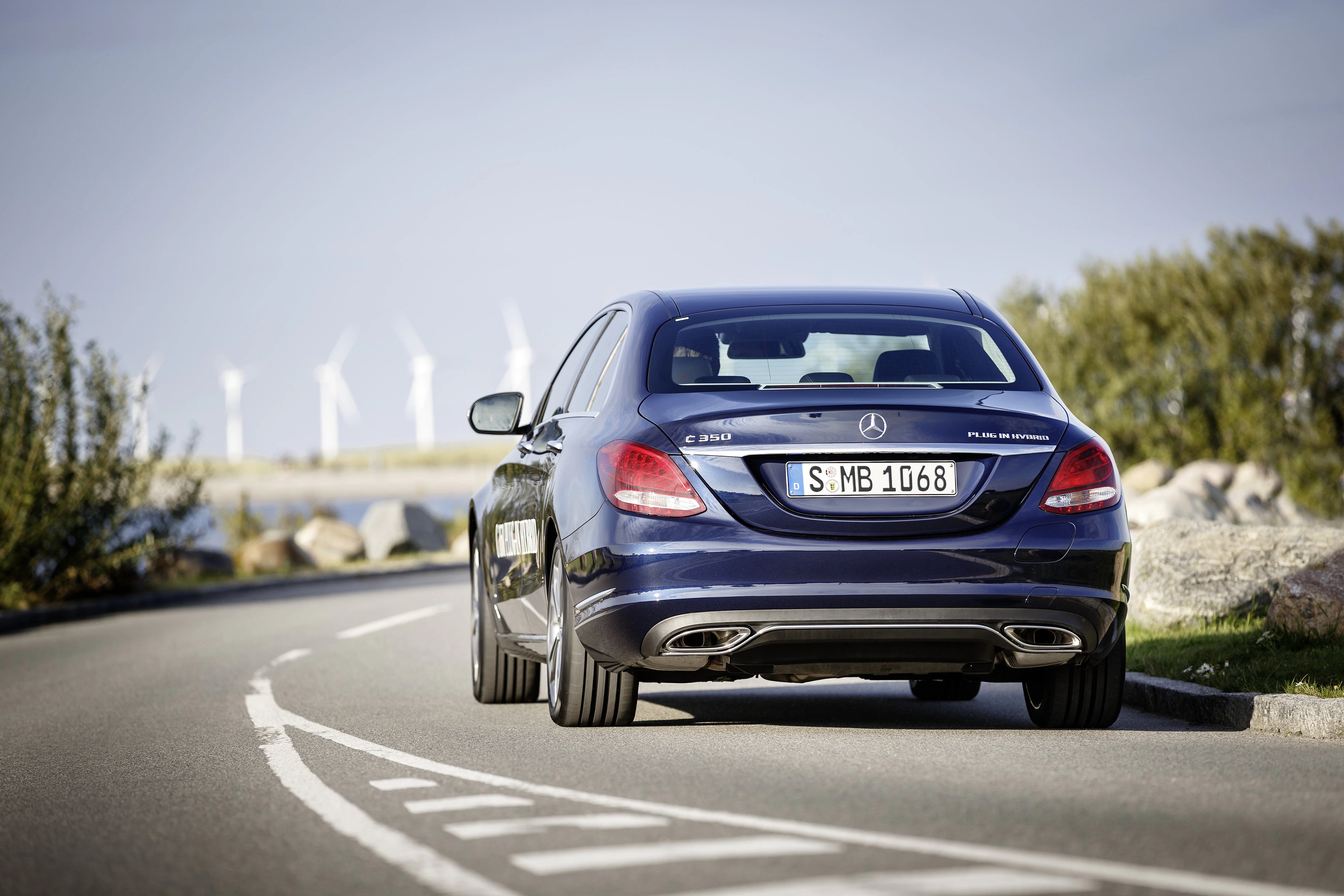 Ein blauer Mercedes-Benz fährt auf einer kurvigen Straße entlang einer Küstenlandschaft mit Windkraftanlagen im Hintergrund.