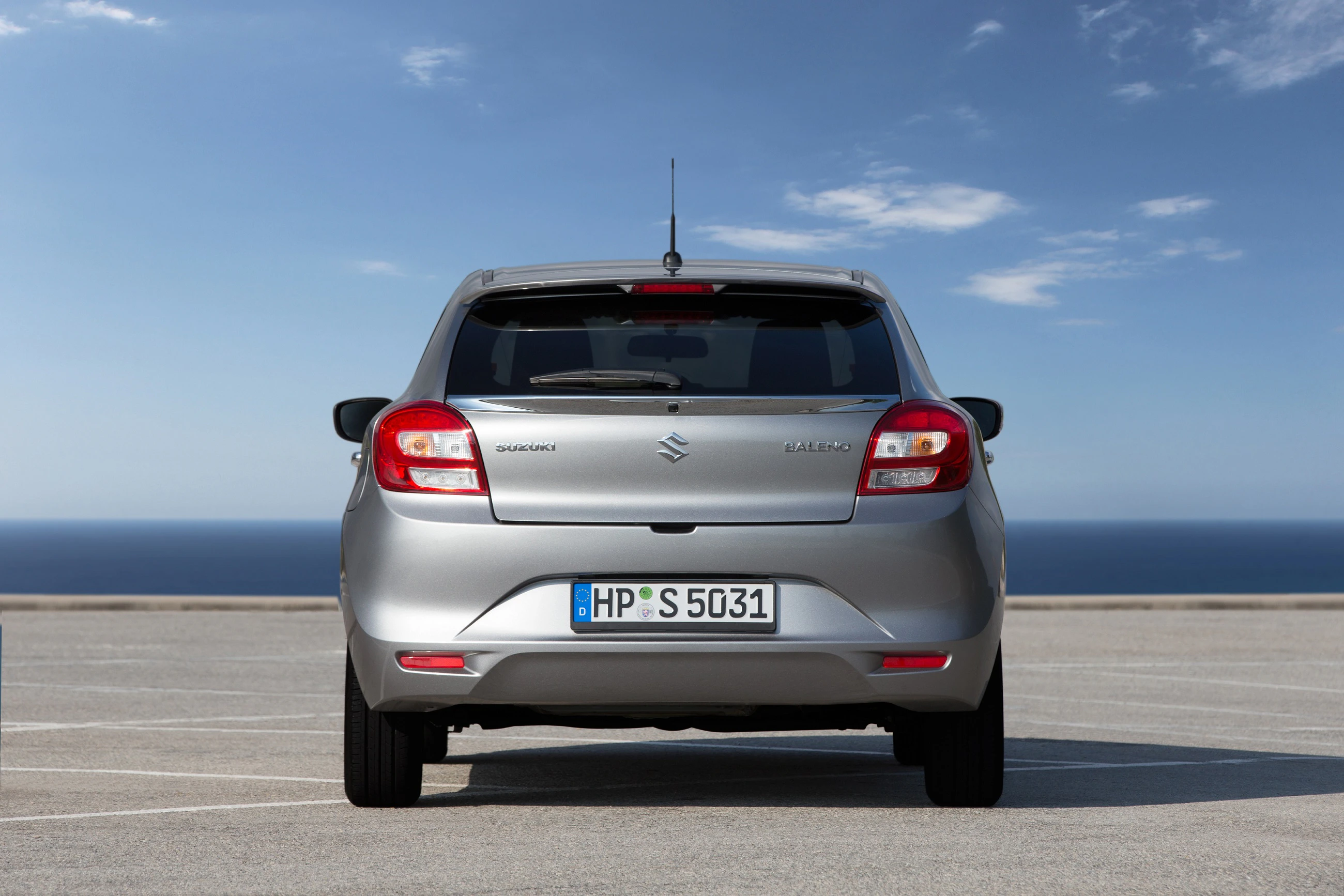 Silberner Suzuki Baleno von hinten auf einem Parkplatz mit Meer und blauem Himmel im Hintergrund.
