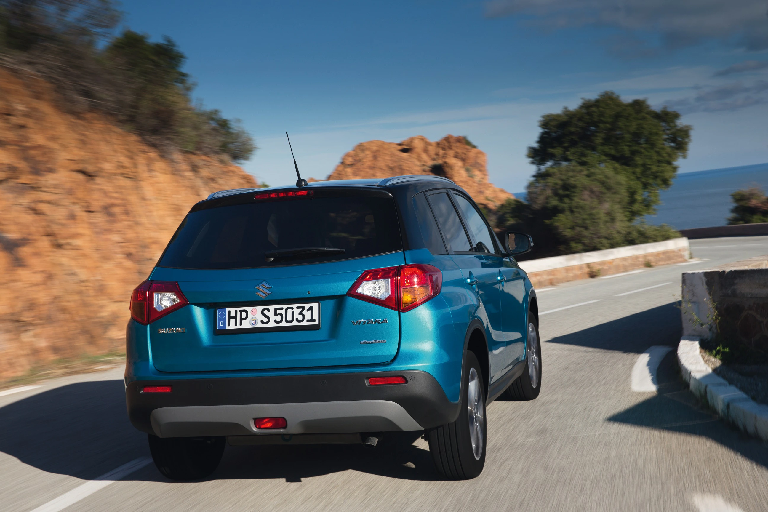 Ein blauer SUV fährt auf einer kurvigen Küstenstraße mit Felsen und Meer im Hintergrund entlang.