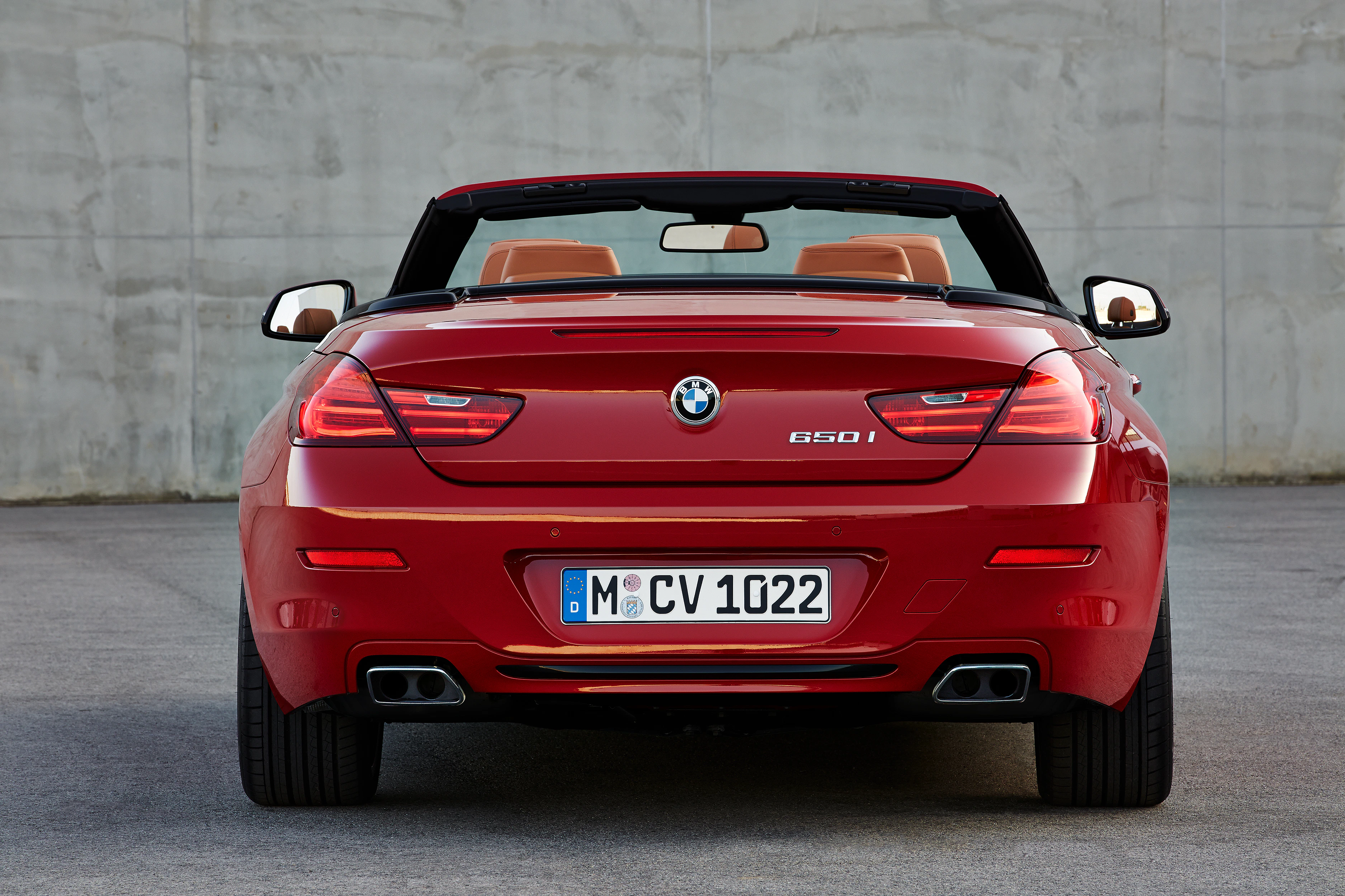 Roter BMW 650i Cabrio von hinten, schwarzes Verdeck geöffnet, auf Asphalt vor grauer Betonwand.