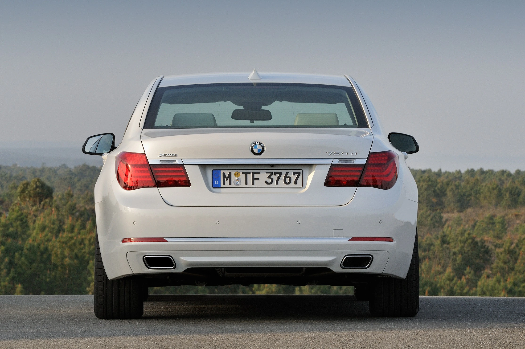 Rückansicht eines weißen BMW 760Li, geparkt auf einer Straße. Im Hintergrund ist bewaldetes Gelände zu sehen.