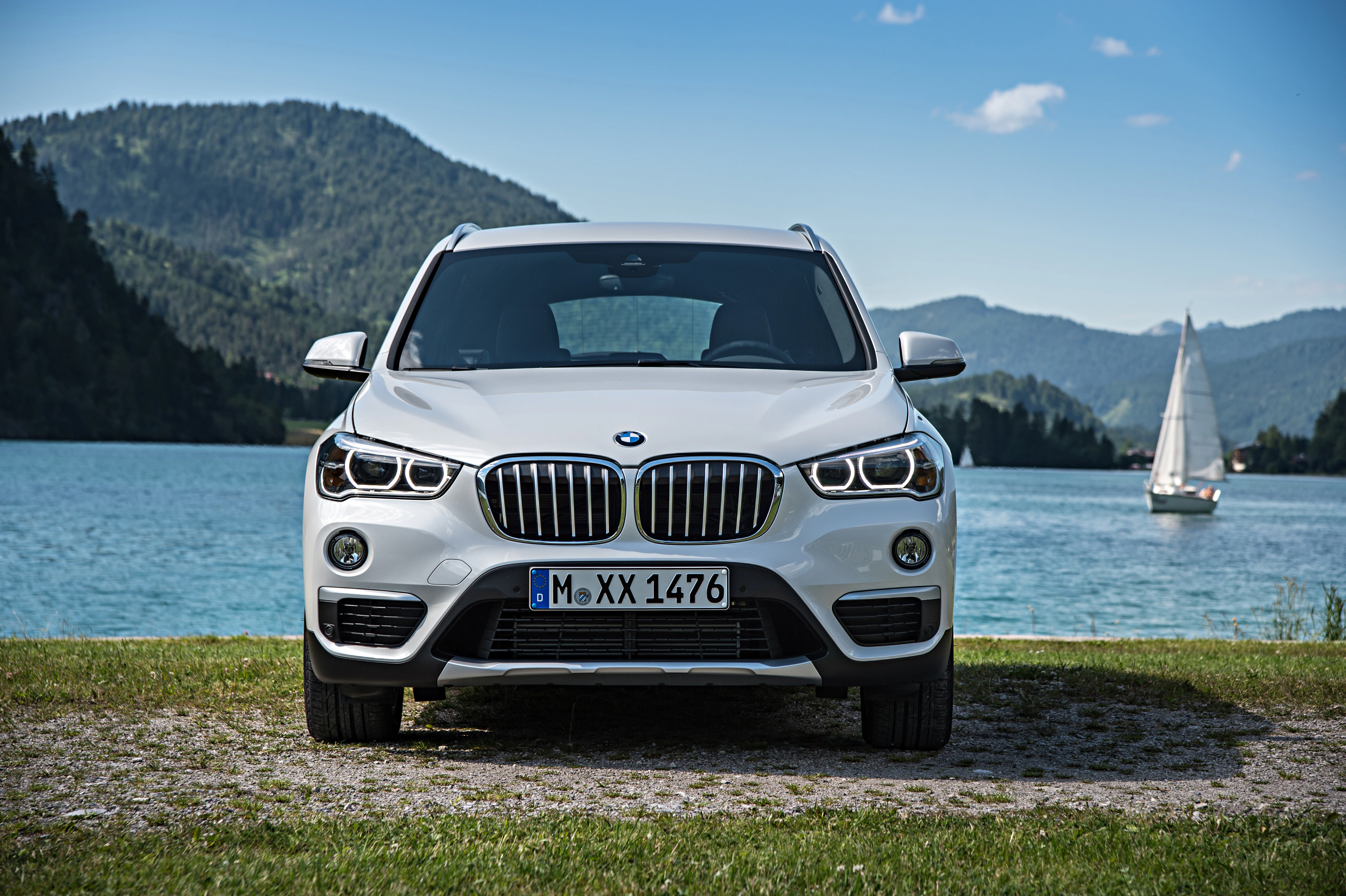 Weißer BMW SUV steht vor einem See mit einem Segelboot im Hintergrund, umgeben von grünen Hügeln und blauem Himmel.