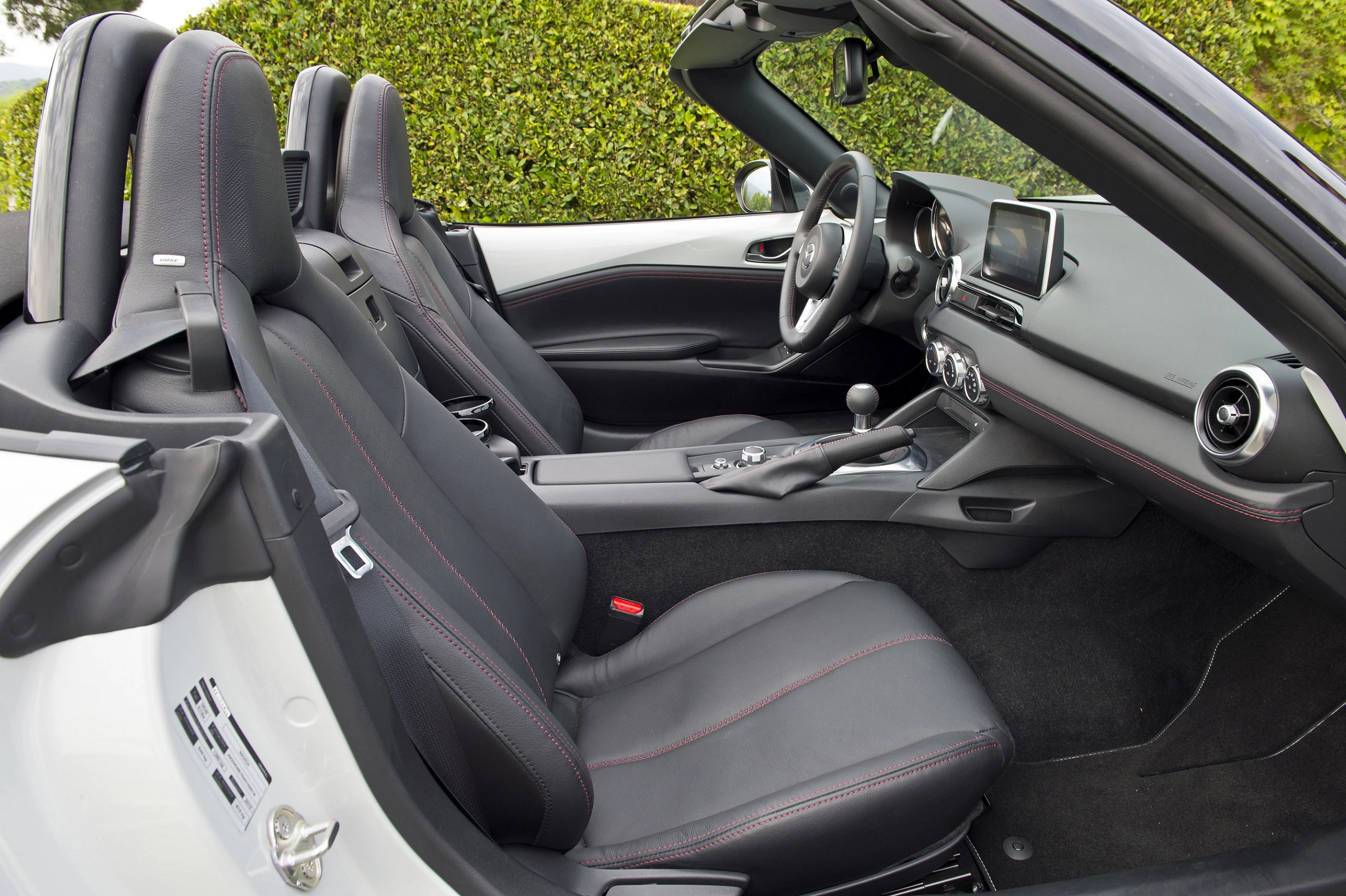 Innenraum eines weißen Cabrios mit schwarzen Ledersitzen und roter Ziernaht. Offenes Verdeck, modernes Armaturenbrett mit Display.
