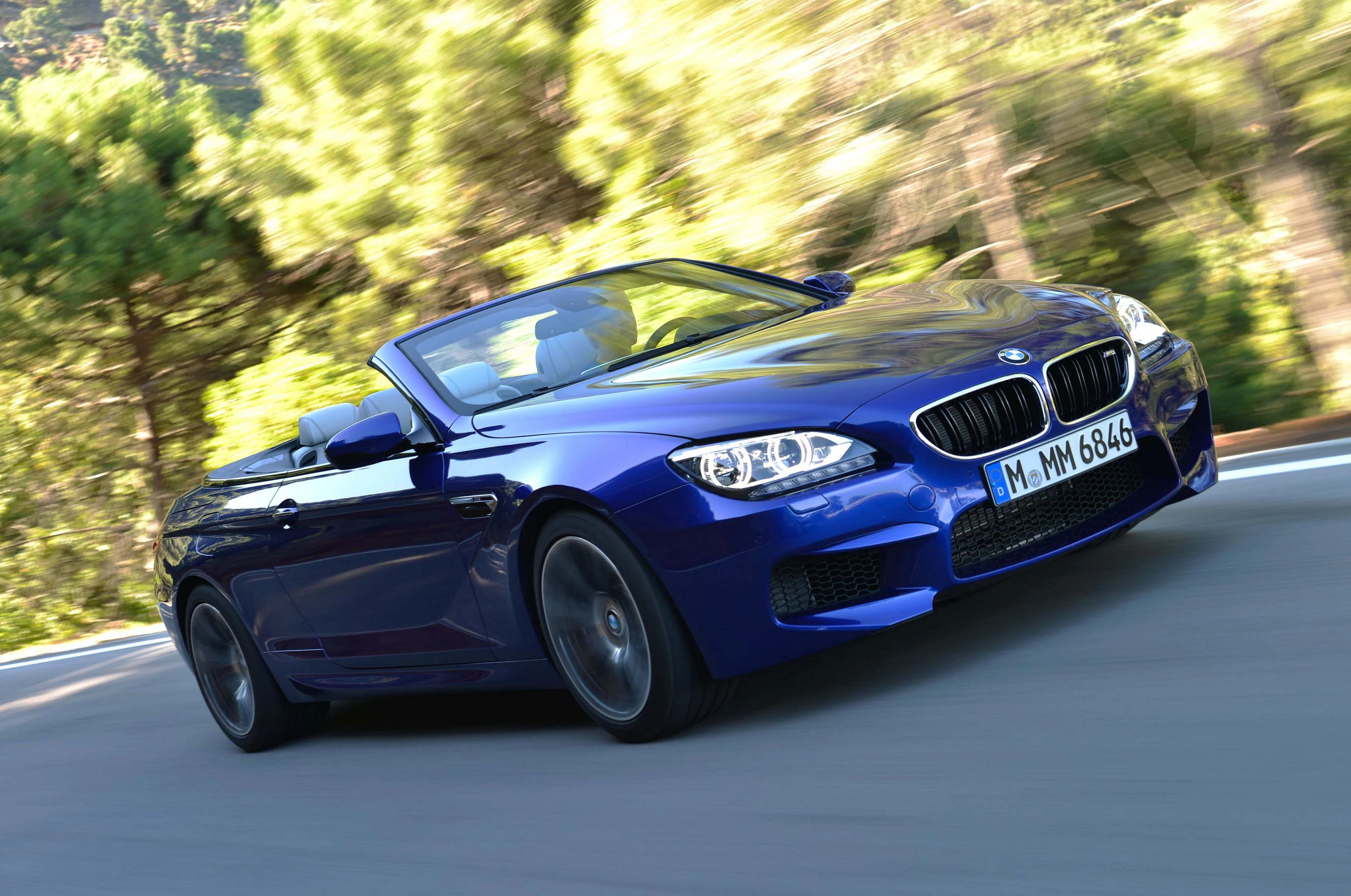 Blaues BMW-Cabrio fährt auf kurviger Landstraße bei sonnigem Wetter. Bäume im Hintergrund.