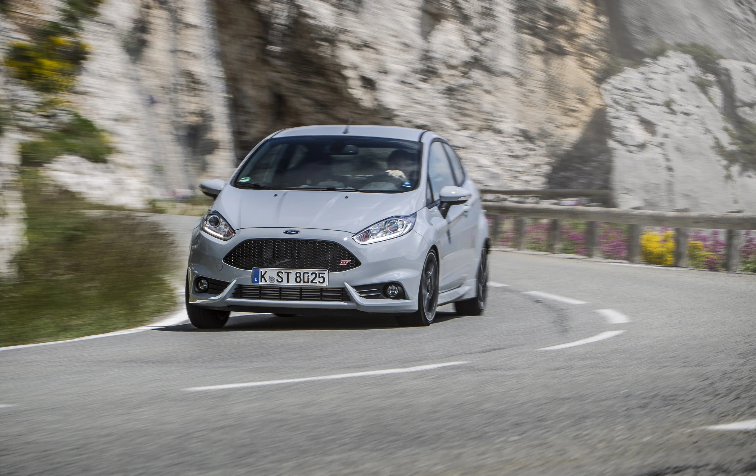 Silberner Ford Fiesta ST fährt auf kurviger Bergstraße, umgeben von Felsen und blühenden Pflanzen.