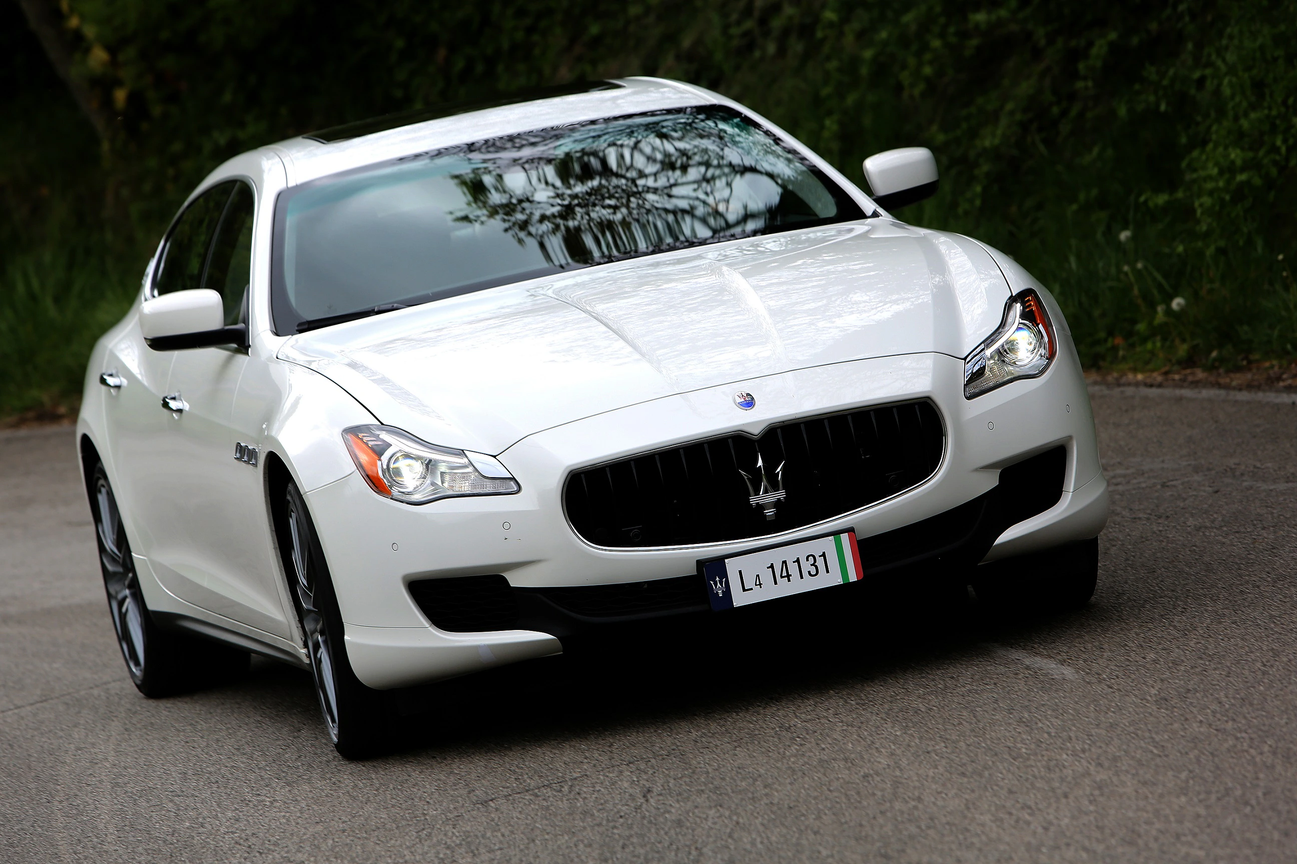 Weißer Maserati Quattroporte fährt auf einer kurvigen Landstraße, mit Bäumen im Hintergrund.