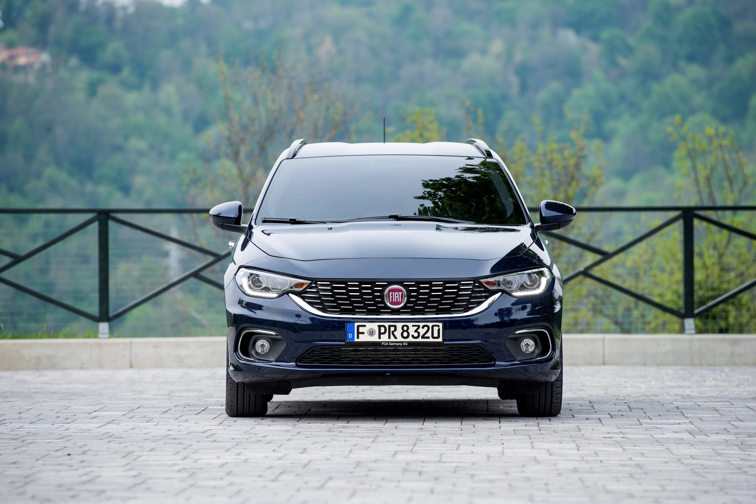 Blauer Fiat Tipo Kombi, frontal fotografiert auf gepflastertem Platz vor grünem Hintergrund. Nummernschild sichtbar.