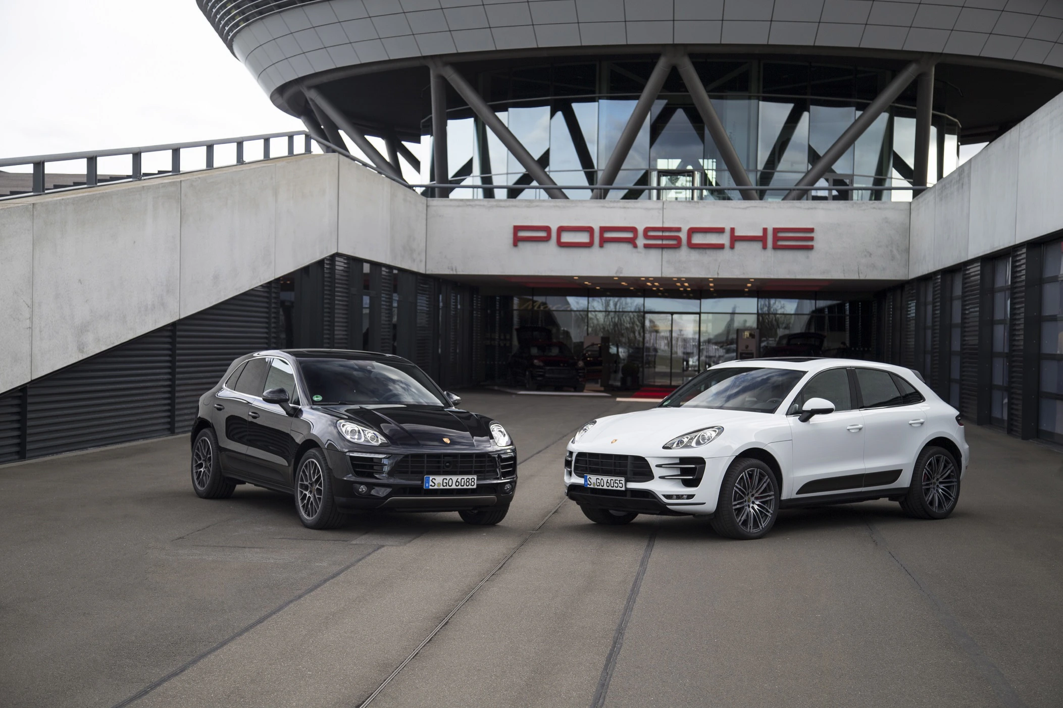Zwei Porsche SUVs, ein schwarzer und ein weißer, parken vor dem Porsche-Gebäude mit großem Logo, moderner Architektur im Hintergrund.